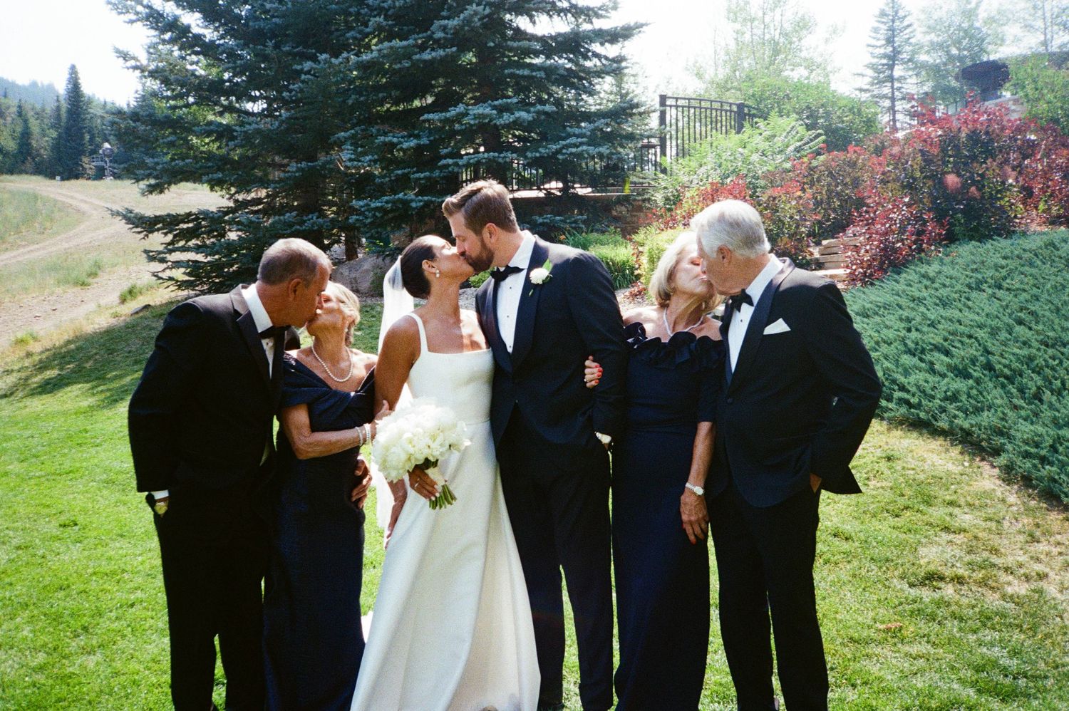 Wedding party members share a joyful moment together on a grassy hillside with evergreen trees in the background.