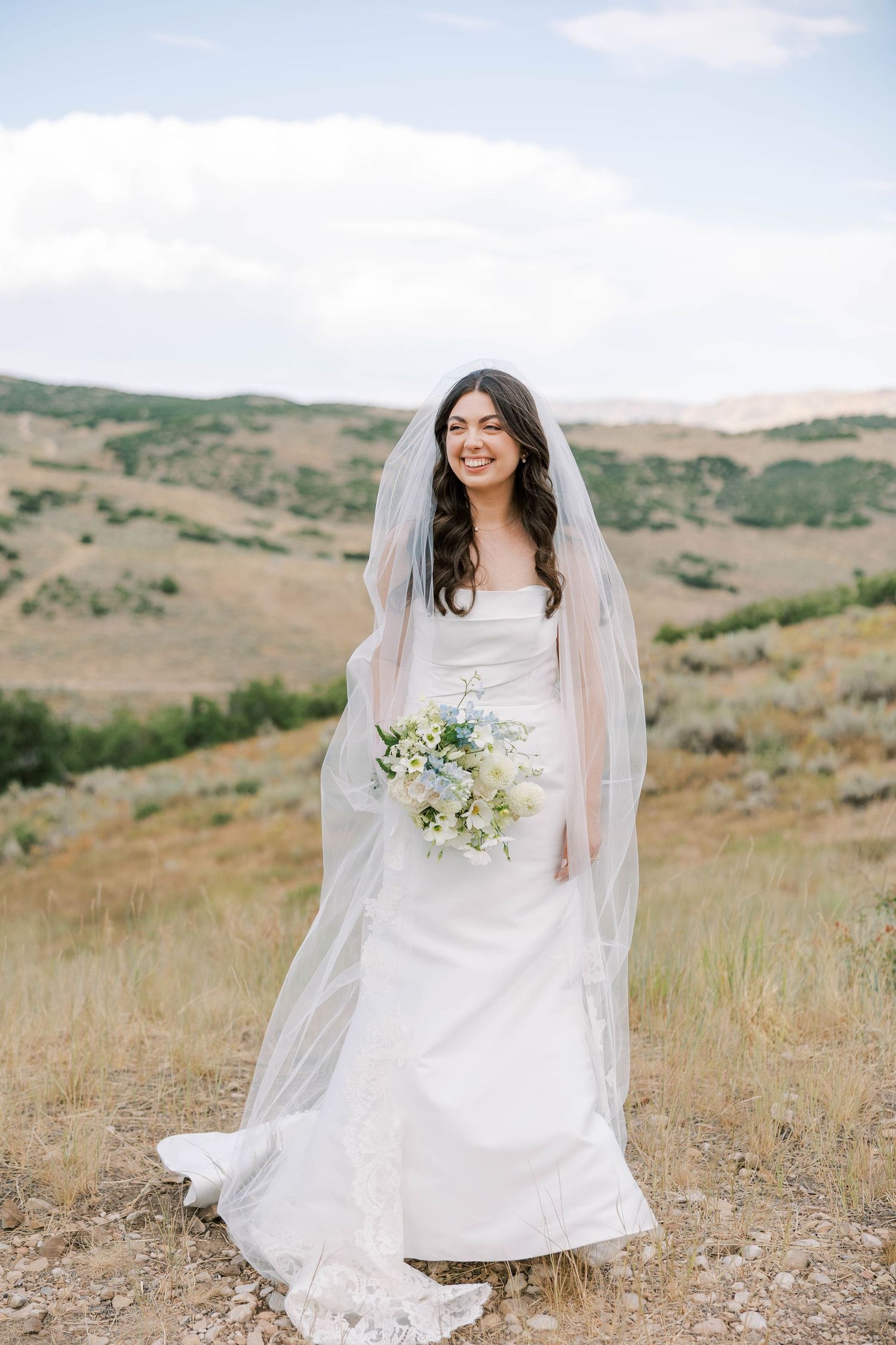 Bride in flowing white dress poses in scenic mountain meadow setting.