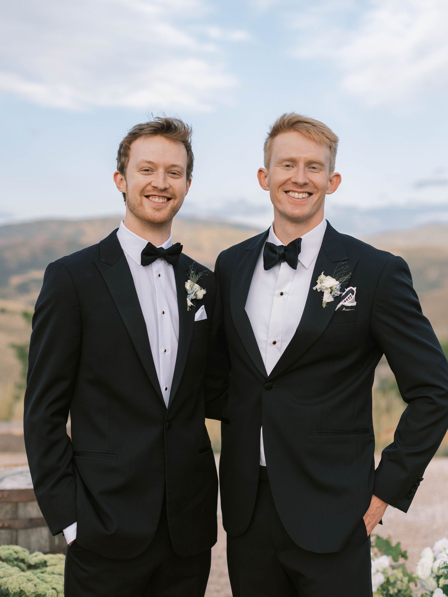 Groomsmen in black tuxedos and bow ties pose together against mountain backdrop at formal wedding.
