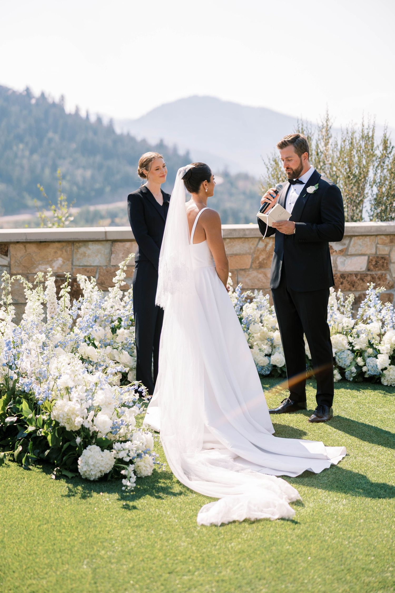 An outdoor wedding ceremony takes place on a terrace overlooking mountains with white floral arrangements and greenery.