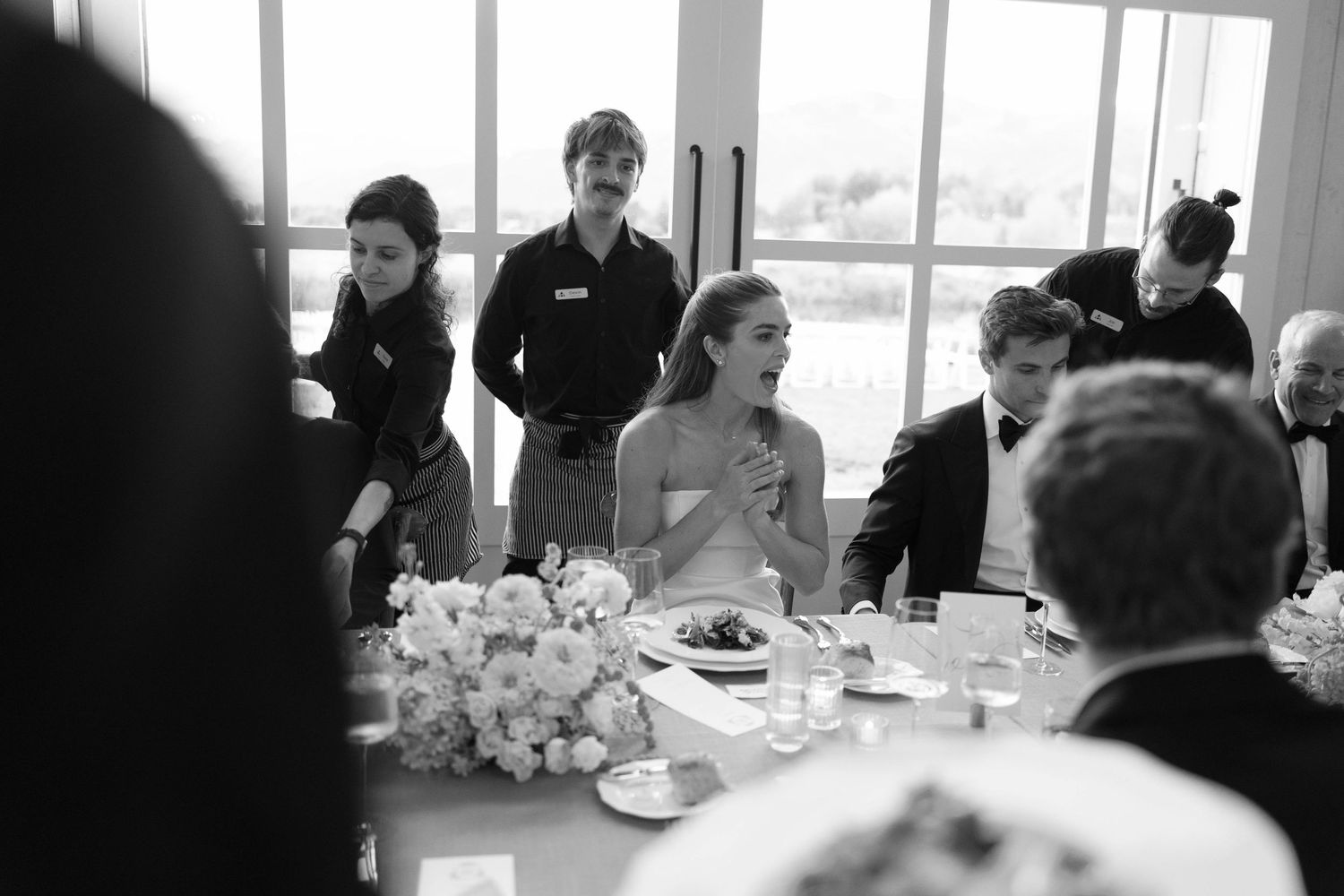 A candid black and white photo of guests at a formal dining event with table settings and floral centerpieces.