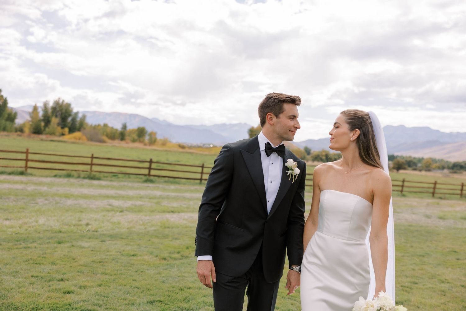 Newlywed couple shares a romantic moment in an open field with mountain views and rustic wooden fences.