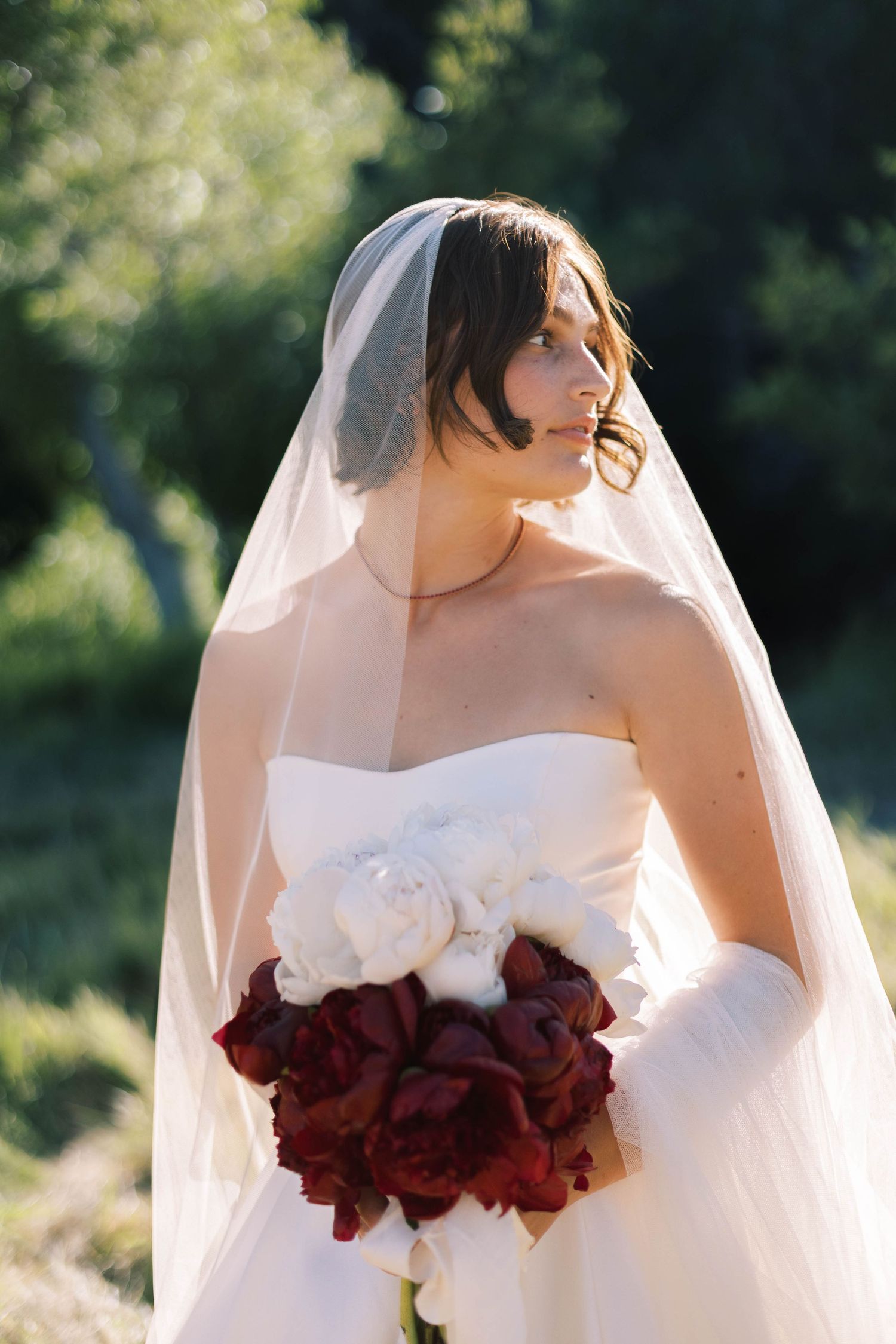 A bride in a white strapless wedding dress holds a deep burgundy rose bouquet in natural outdoor lighting.