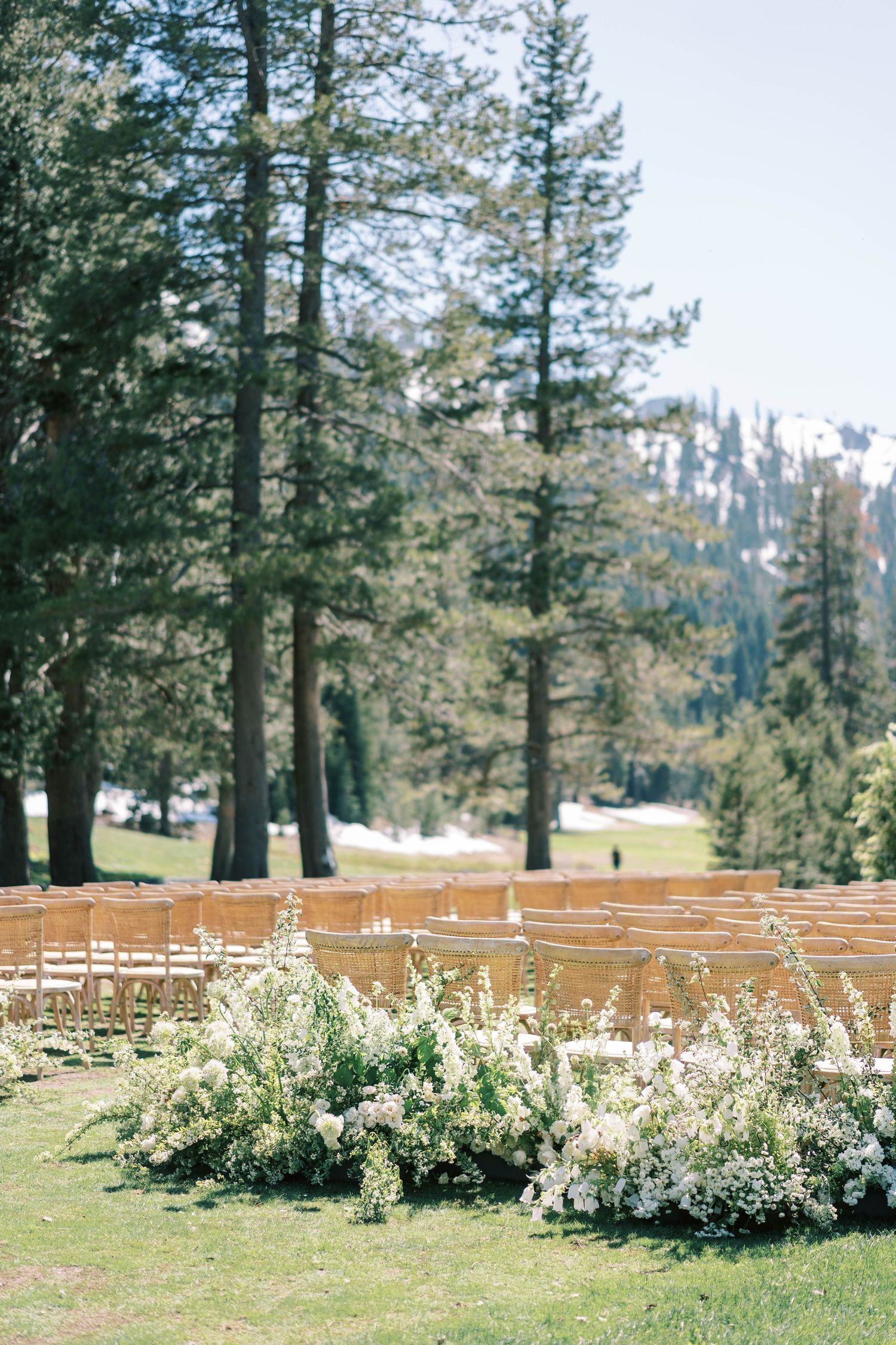Outdoor wedding ceremony setup with white floral arrangements and wooden chairs against mountain scenery.