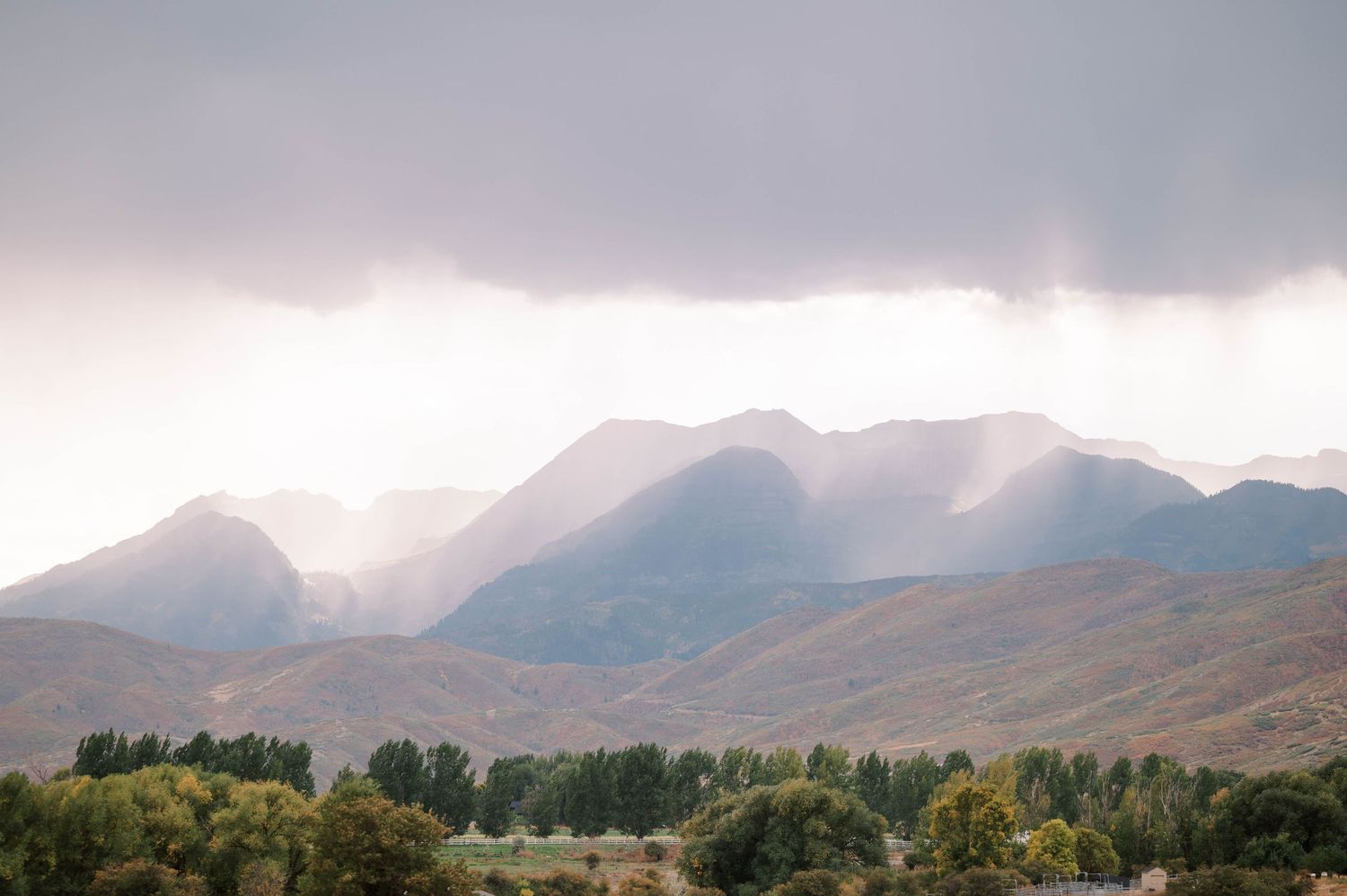 Sunbeams pierce through clouds over rugged mountain peaks with a tree-lined landscape in the foreground.