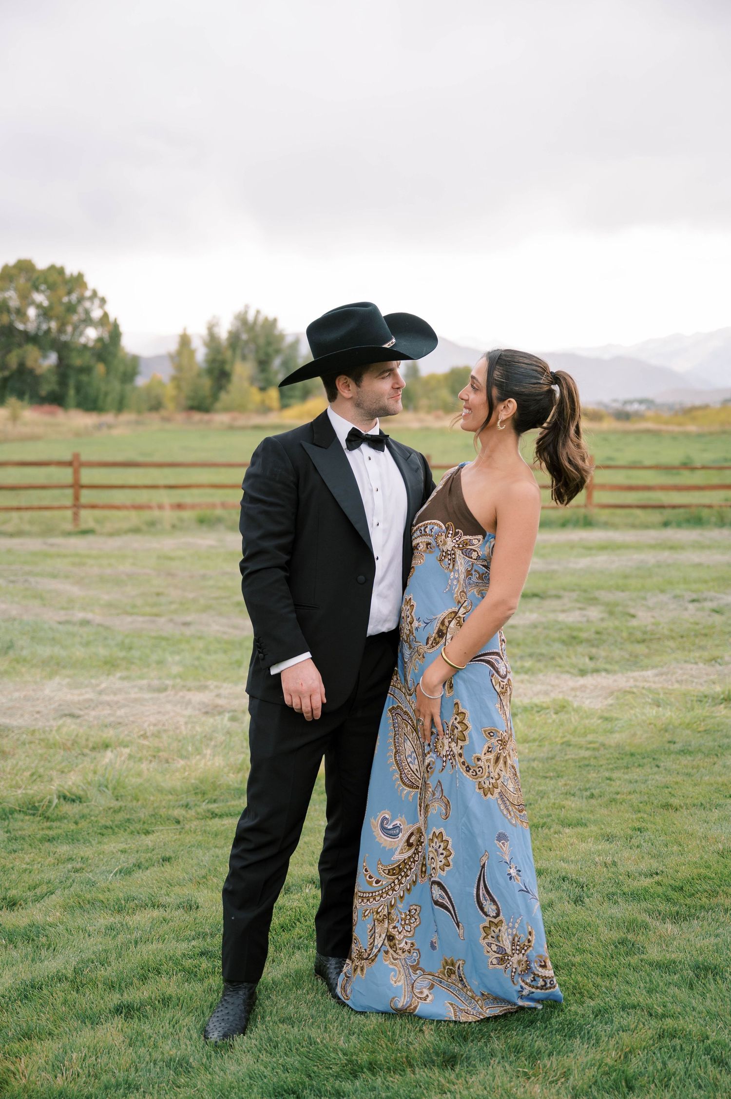 A couple in formal attire share a playful moment in a grassy field with mountains and wooden fencing in the background.