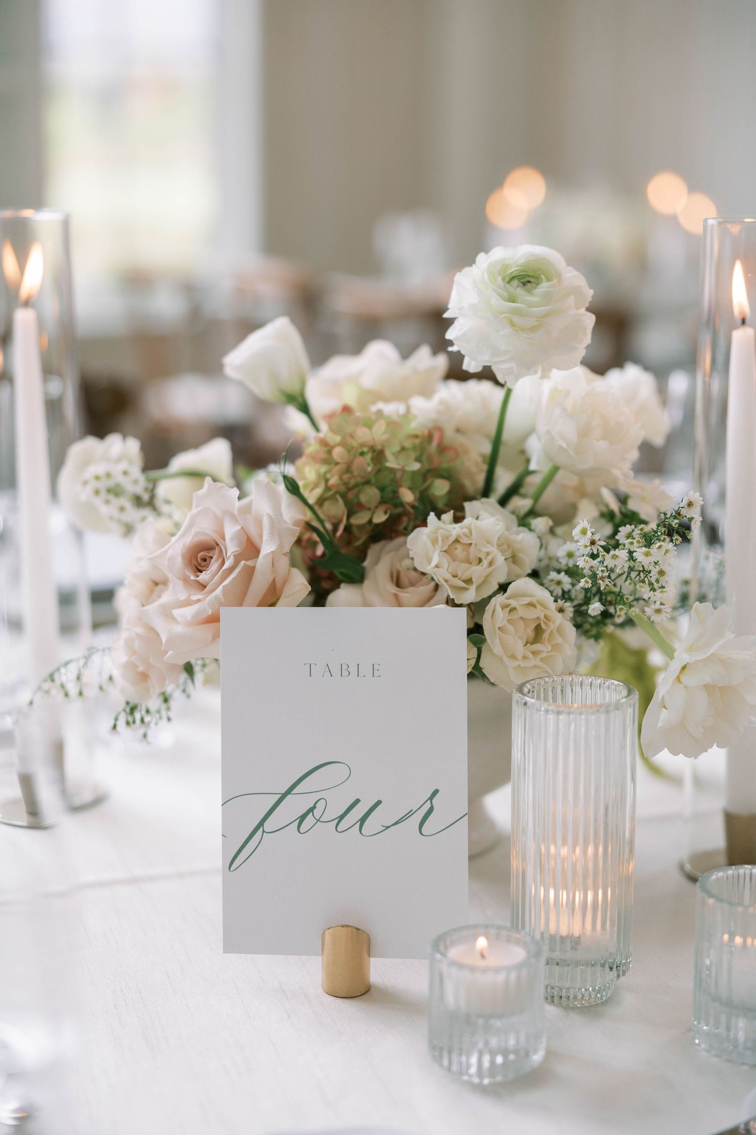 Elegant white floral wedding table centerpiece with candles and calligraphy table number in soft romantic lighting.