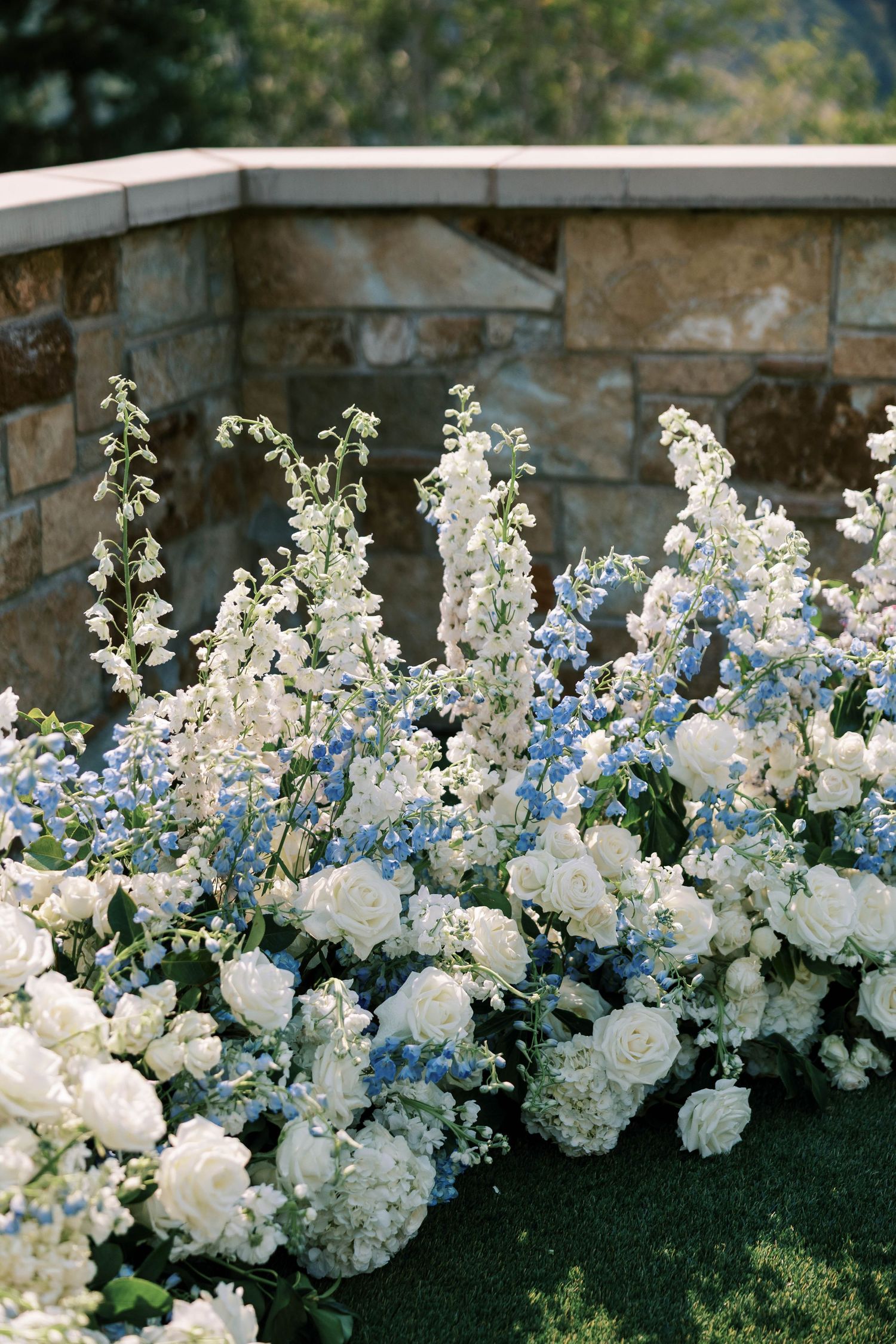 Luxurious white and blue flower arrangements with roses and delphinium create a stunning border along a stone wall.