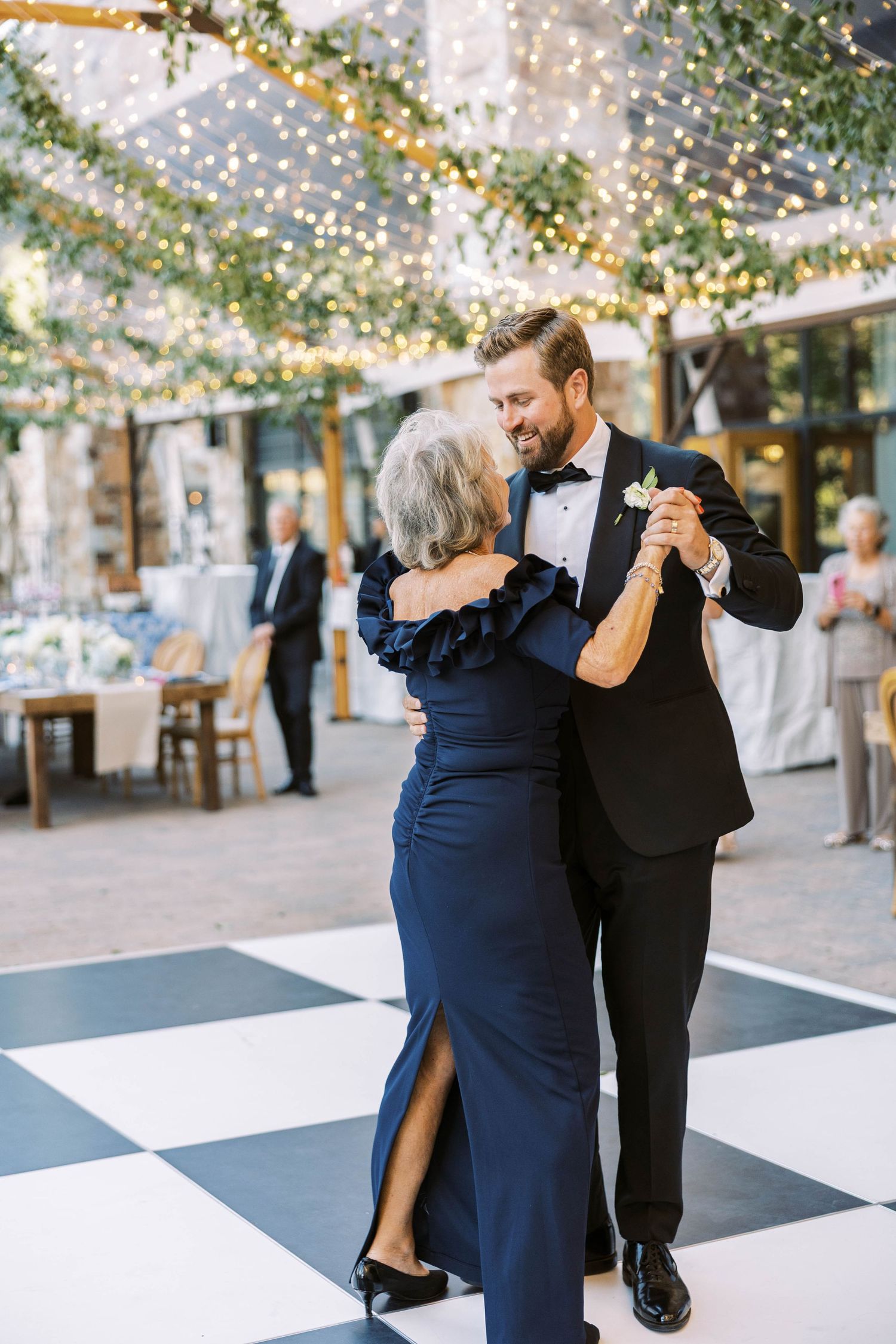 A couple dances elegantly on a black and white checkered floor under twinkling lights at an outdoor venue.