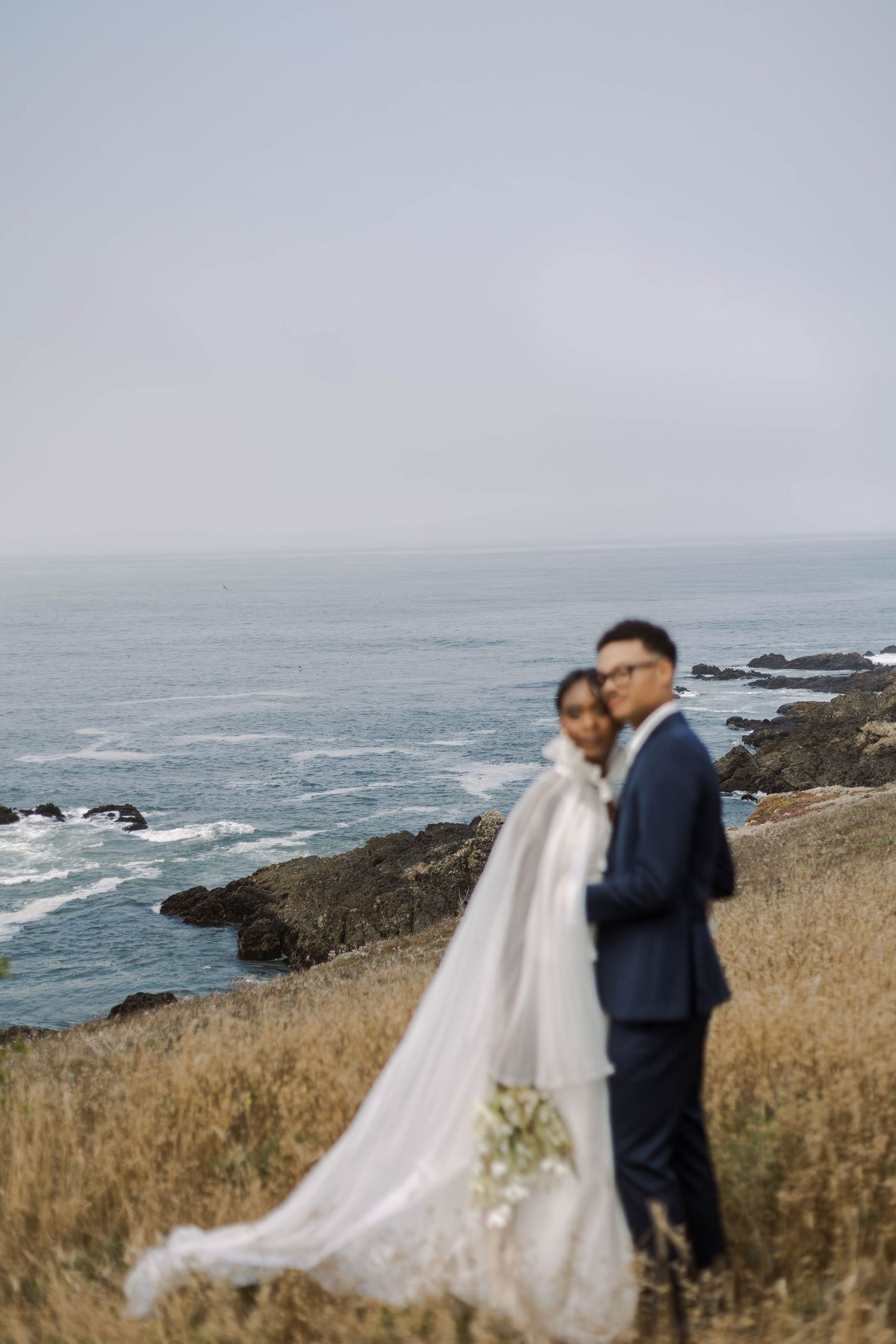 A couple in wedding attire stands on a scenic coastal cliff overlooking the ocean and rocky shoreline.