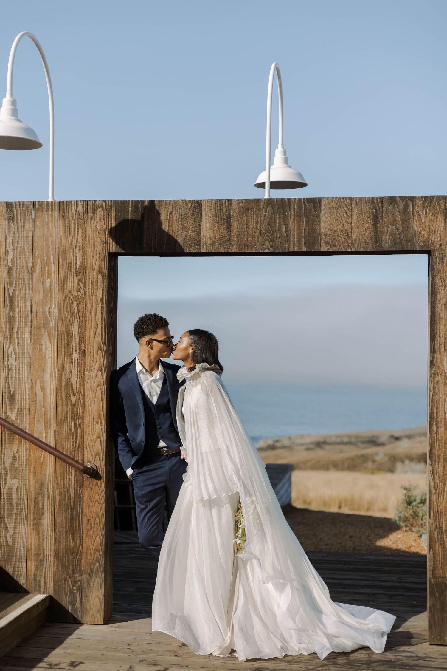 Romantic wedding couple embracing on wooden deck with ocean view and white pendant lights overhead.