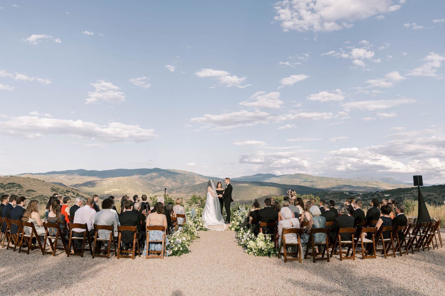A panoramic view of an outdoor wedding ceremony in a scenic mountain landscape with guests seated on wooden chairs.