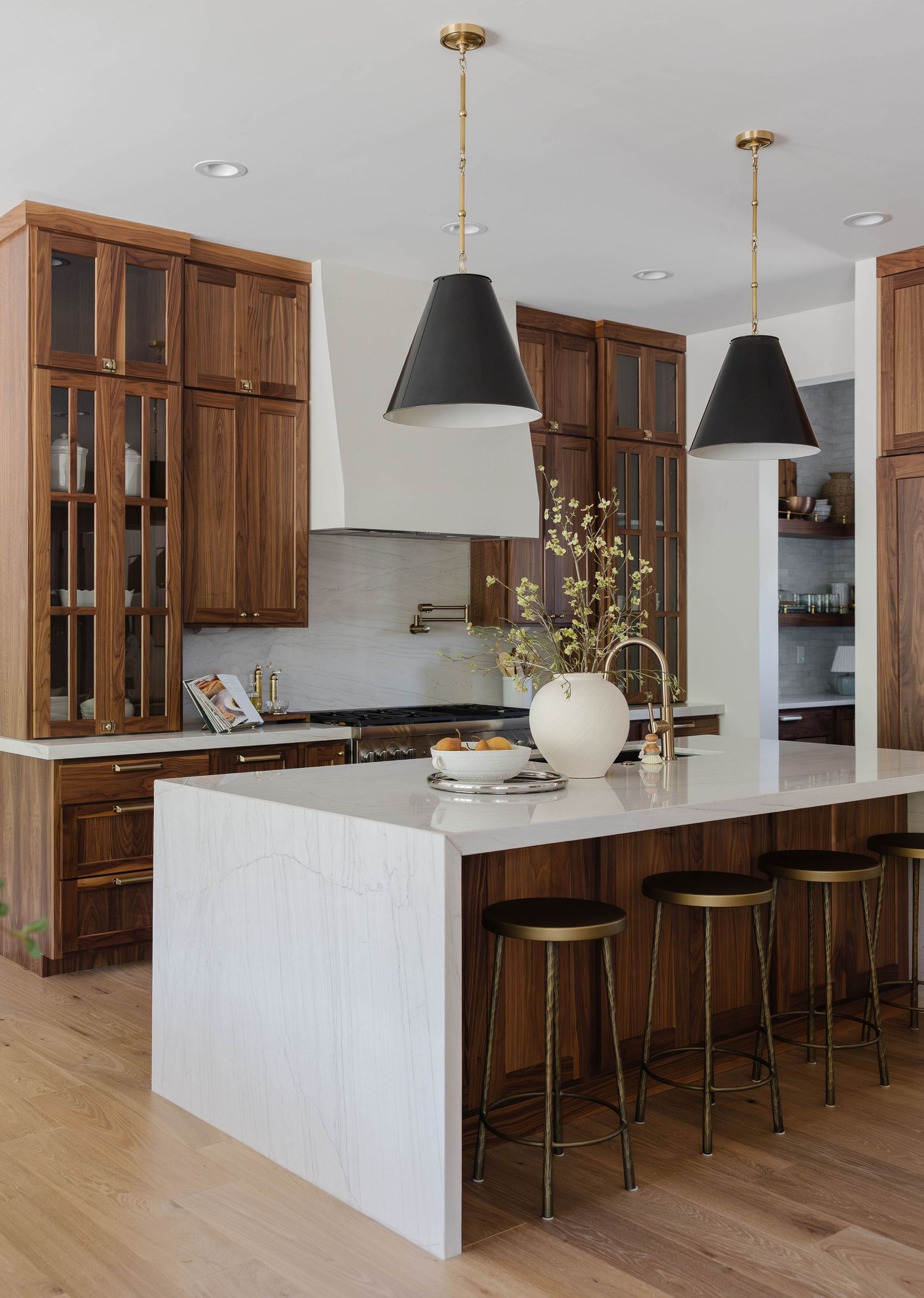 Modern kitchen with dark wood cabinets, white marble island, brass bar stools, and black cone pendant lights on light hardwood floors.