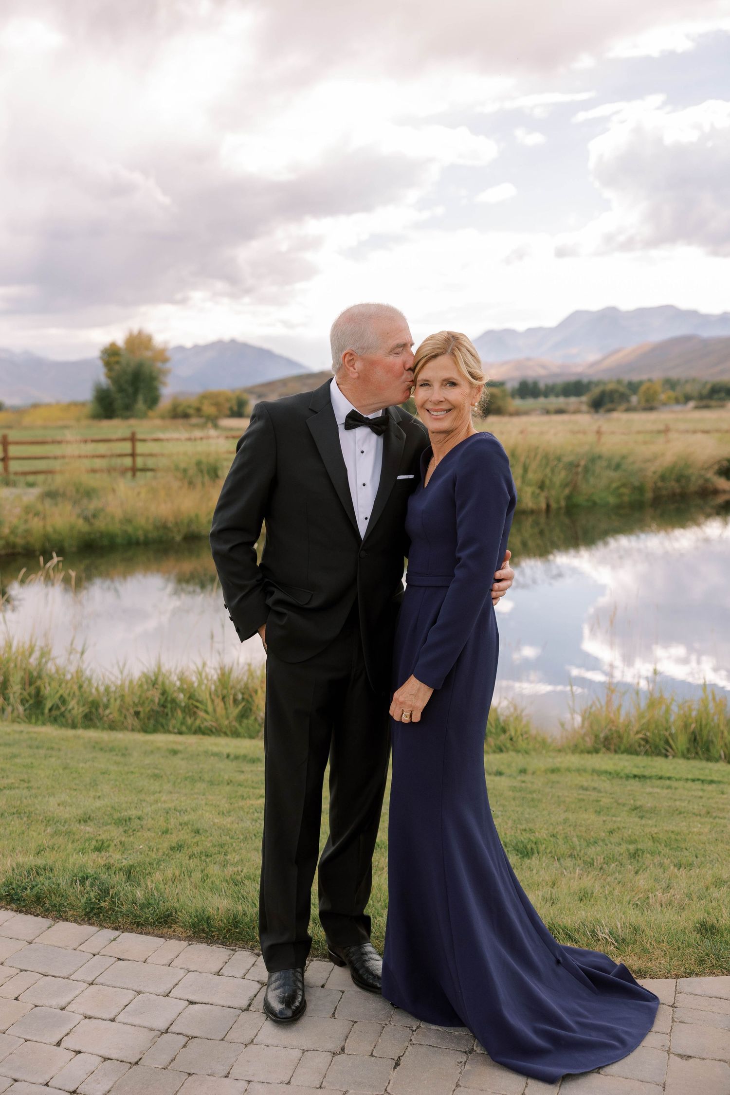 A formal couple in black tie and navy blue gown pose by a pond with mountains in the background.