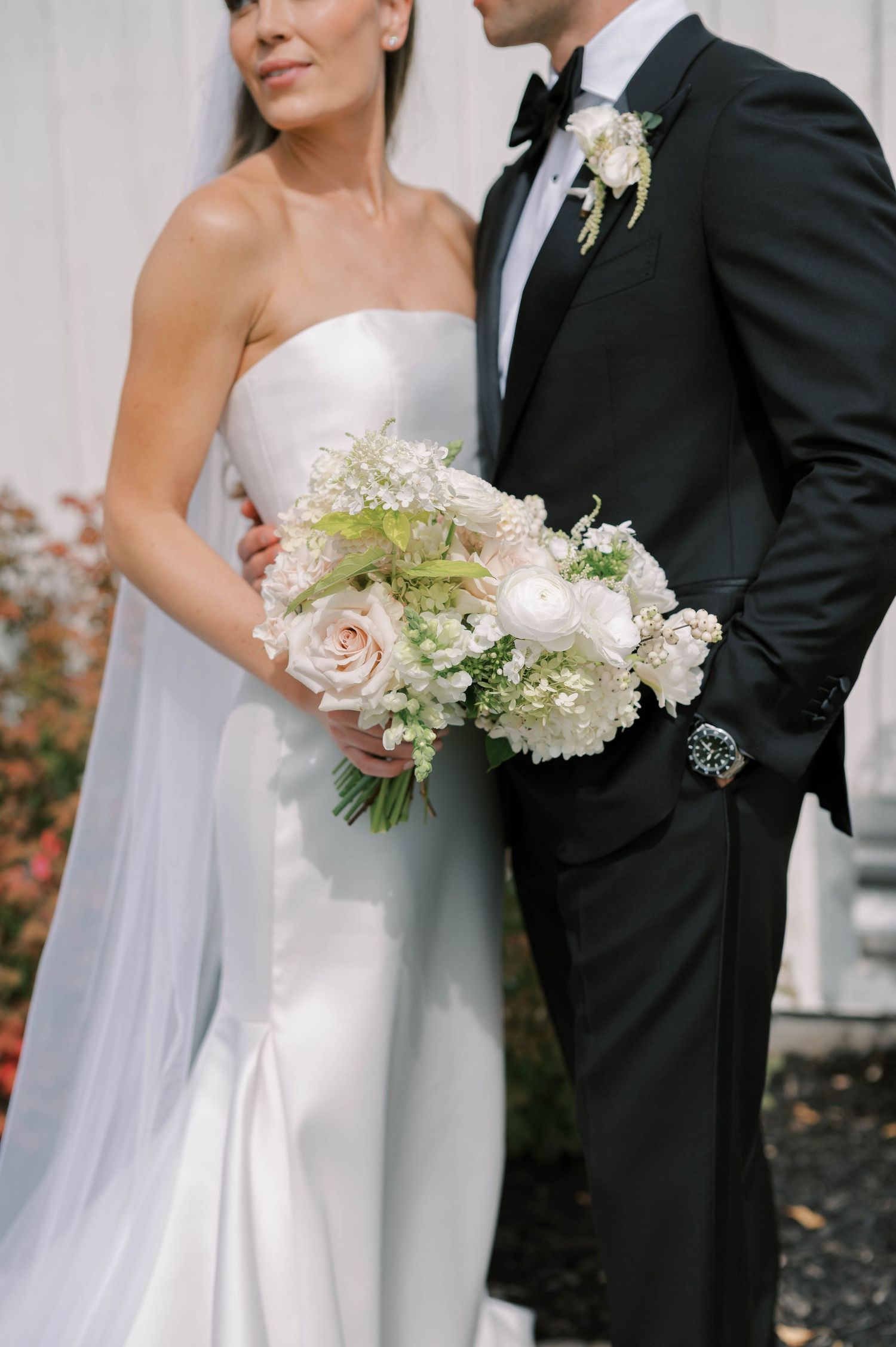 Elegant wedding couple holding white and green floral bouquet against white backdrop.