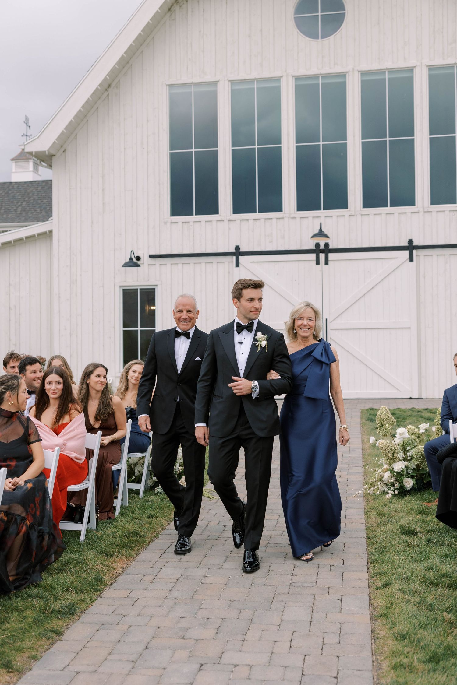Wedding party members walk in a line along a stone path outside a white barn venue with large windows.