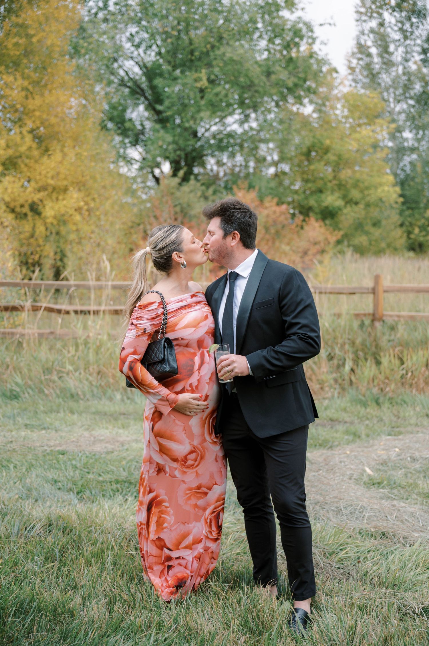 A couple in formal attire shares a romantic moment outdoors on a fall day with golden trees in the background.