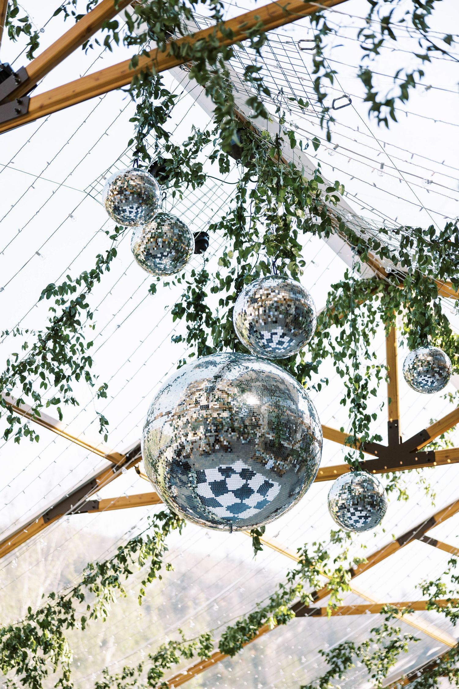 Shimmering disco balls and greenery hang from wooden beams in an elegant tent or canopy setting.