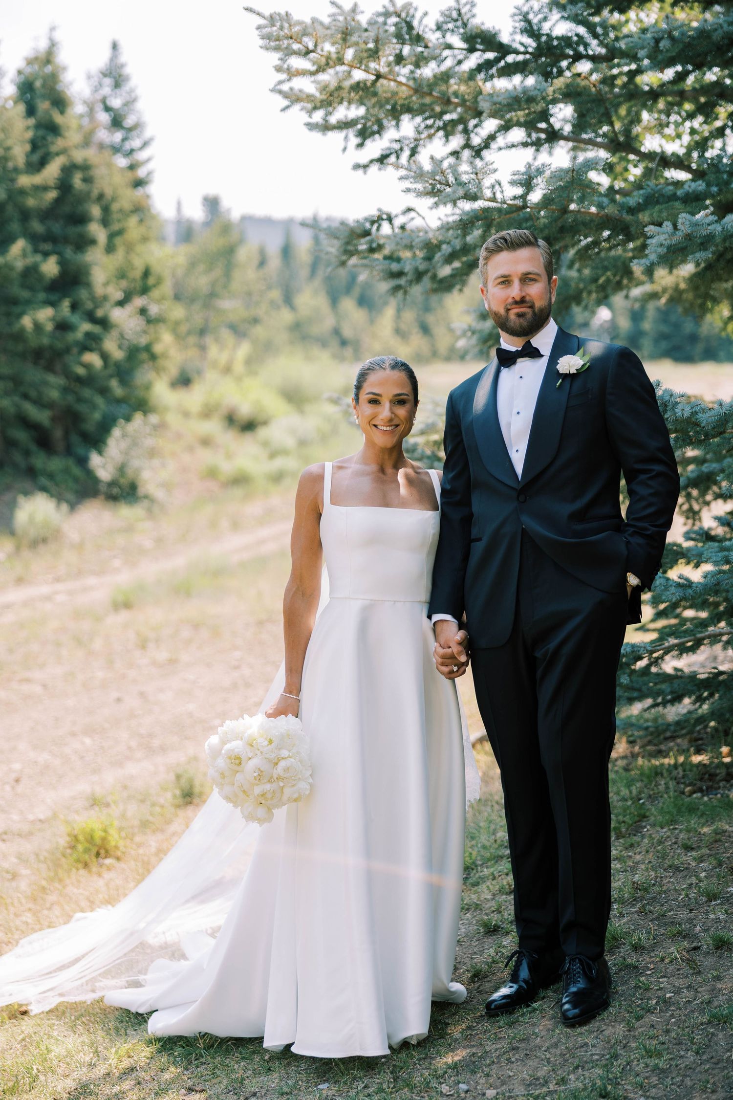 A wedding couple poses together outdoors among evergreen trees on a hillside.