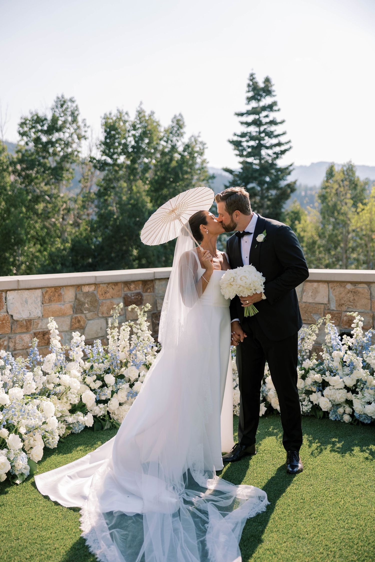 A romantic wedding scene unfolds on a stone terrace with white floral arrangements and mountain views in the background.