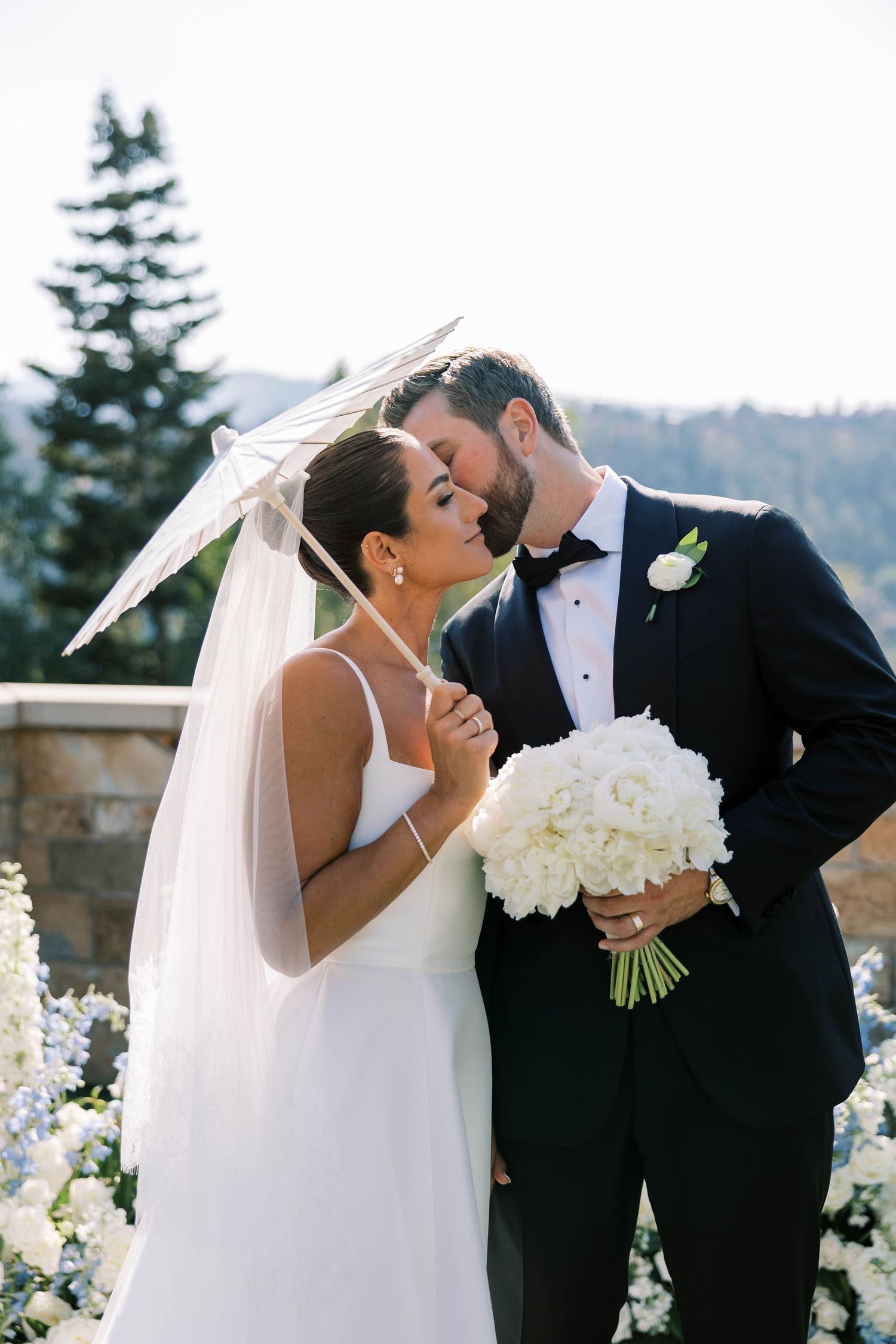 A romantic wedding scene with a white parasol and bouquet of white roses against a mountain backdrop.