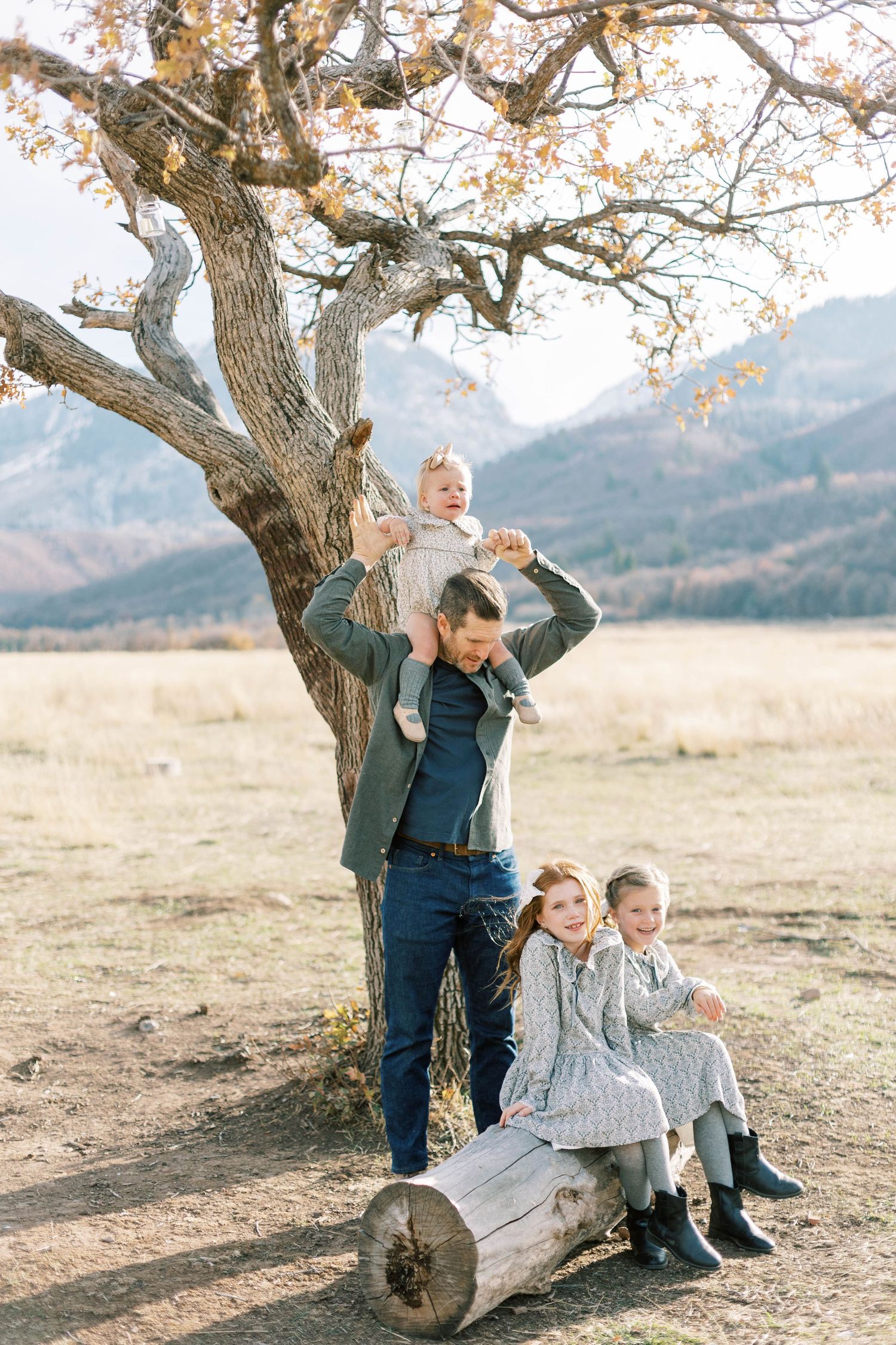 Playful family moment under twisted tree with children sitting on log and mountains behind.