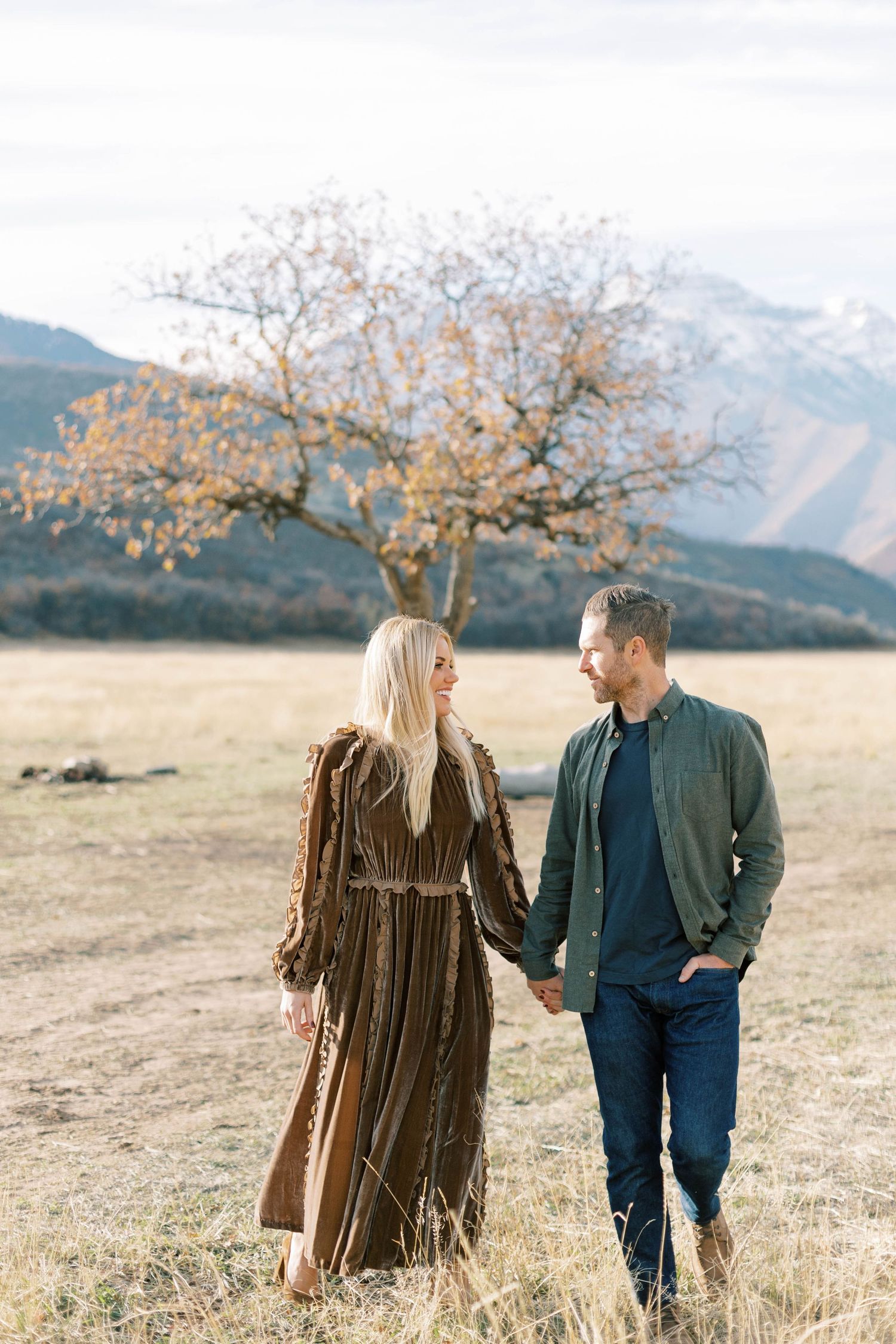 Couple in casual and vintage attire walking through mountain meadow holding hands.
