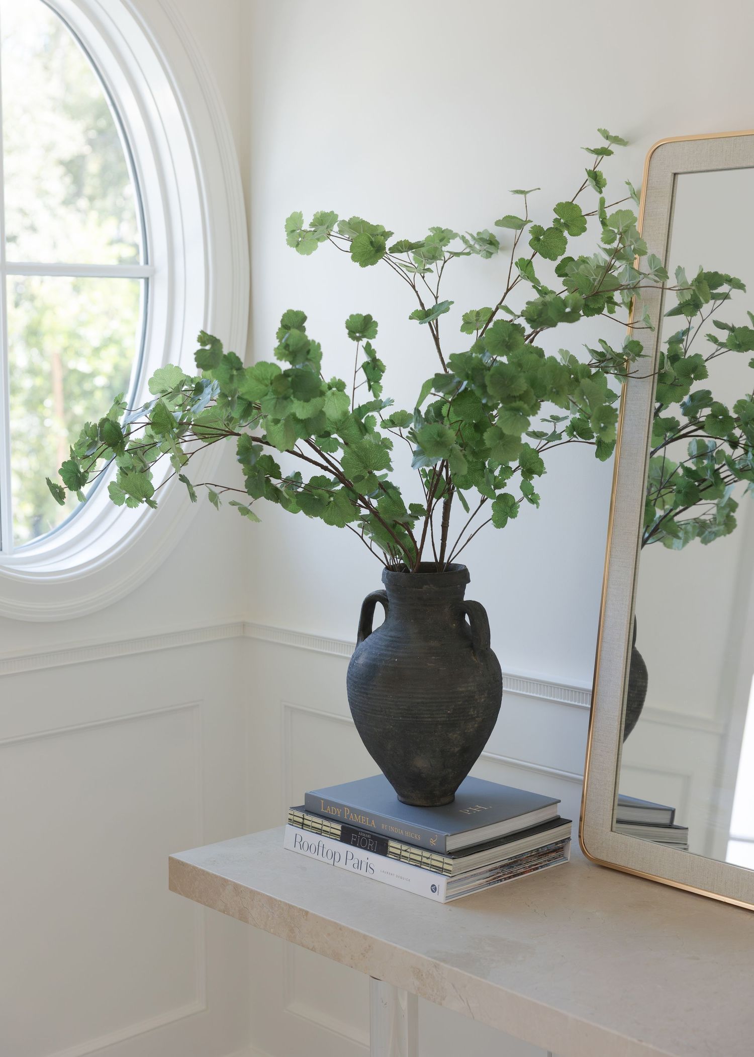 Dark ceramic vase with green eucalyptus branches on stacked books beside a mirror and oval window.