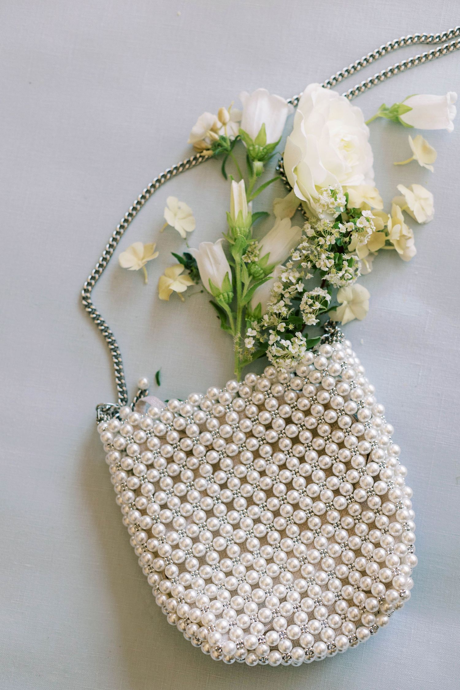 Pearl-embellished bridal purse with white flowers and delicate greenery on gray background.