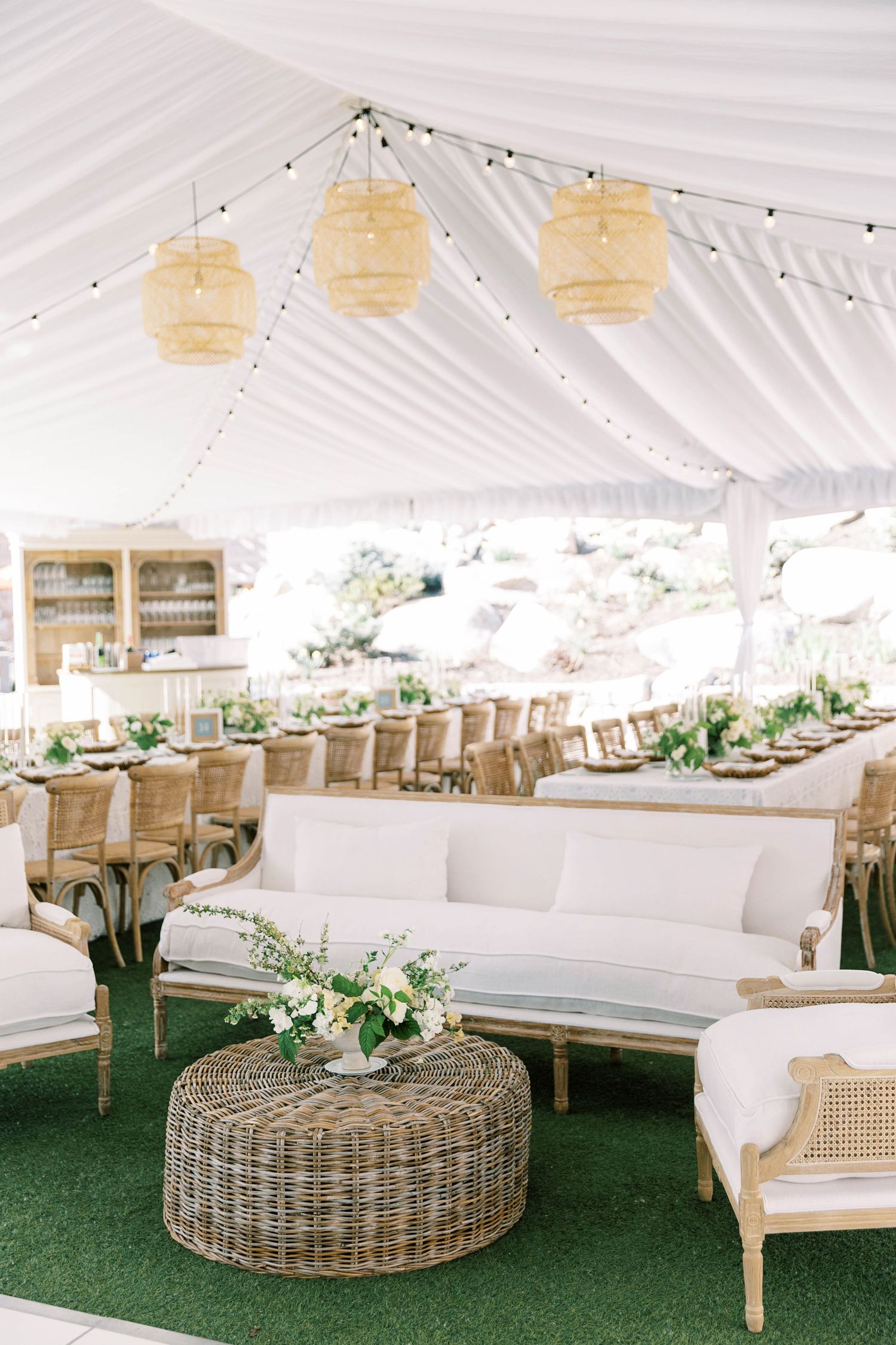 Elegant outdoor wedding tent setup with rattan furniture, white draping, and pendant lanterns creating a bohemian atmosphere.