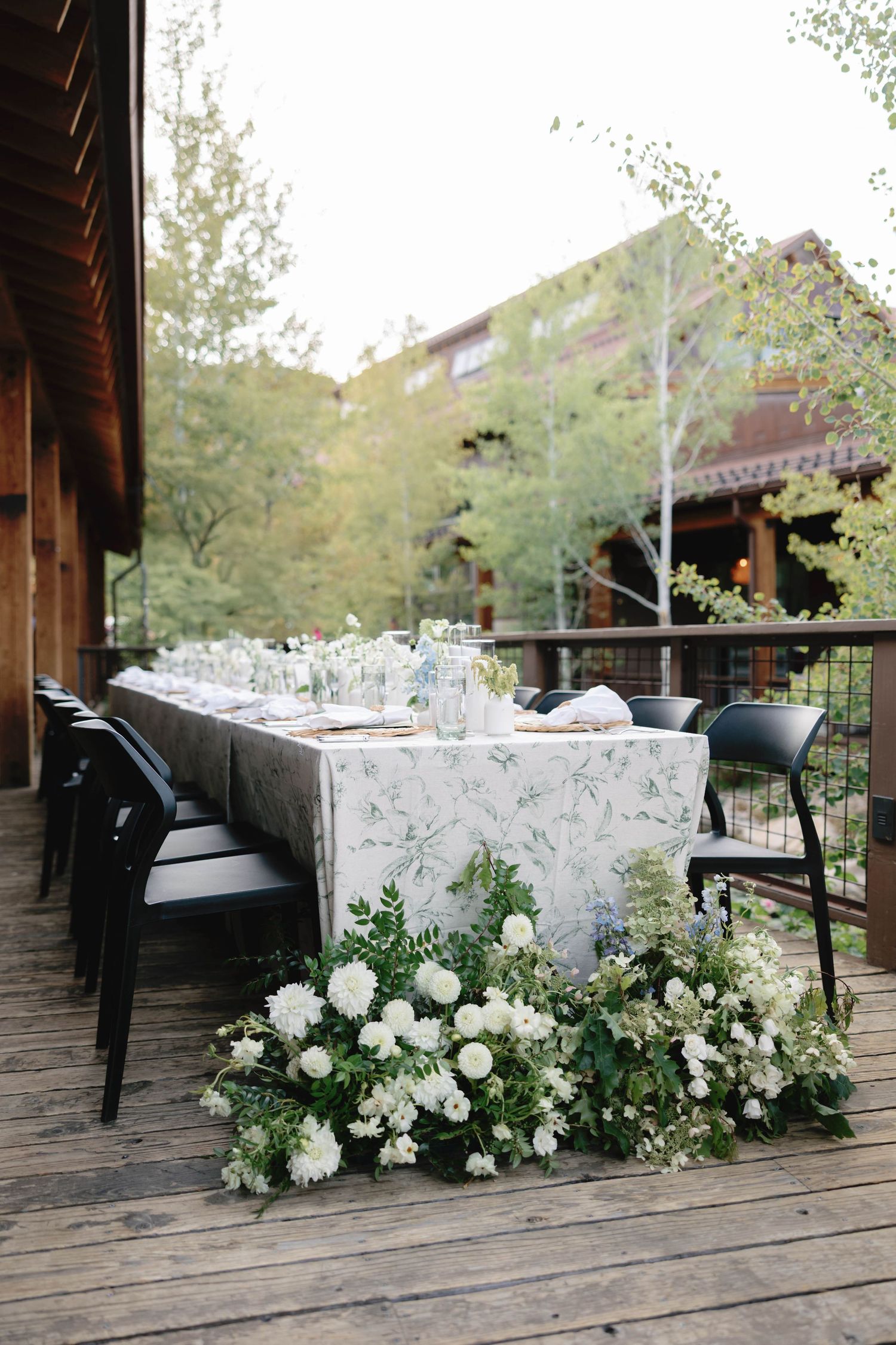 A long dining table with white floral arrangements set on a wooden deck with mountain views.