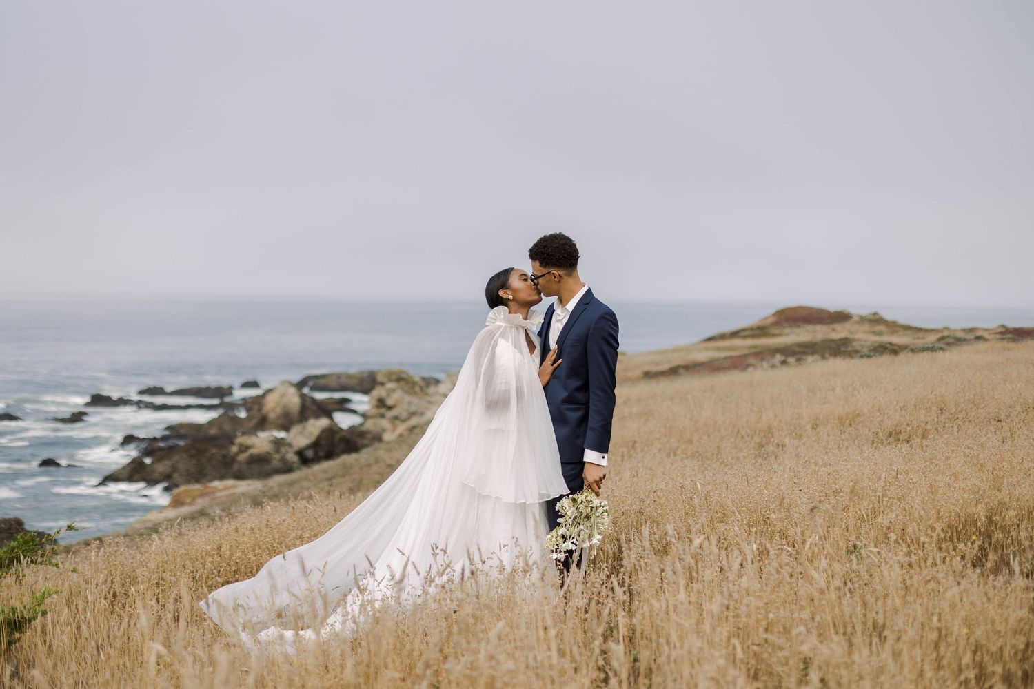 Couple in wedding attire embracing in a golden field overlooking the rocky coastline on a cloudy day.