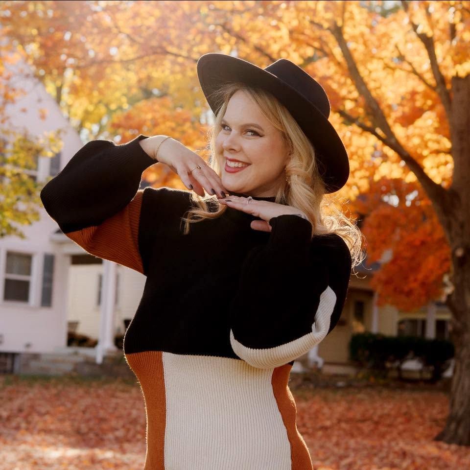 Blonde woman in black hat and colorful sweater posing outdoors among vibrant autumn foliage.