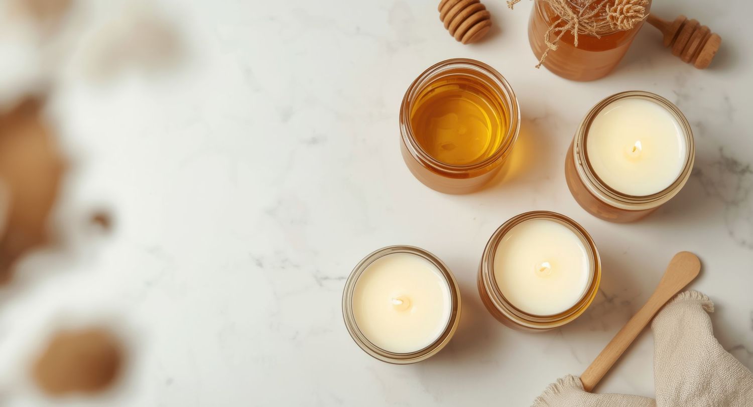 Flat lay of white wax candles in glass jars with honey and wooden dipper on marble surface.