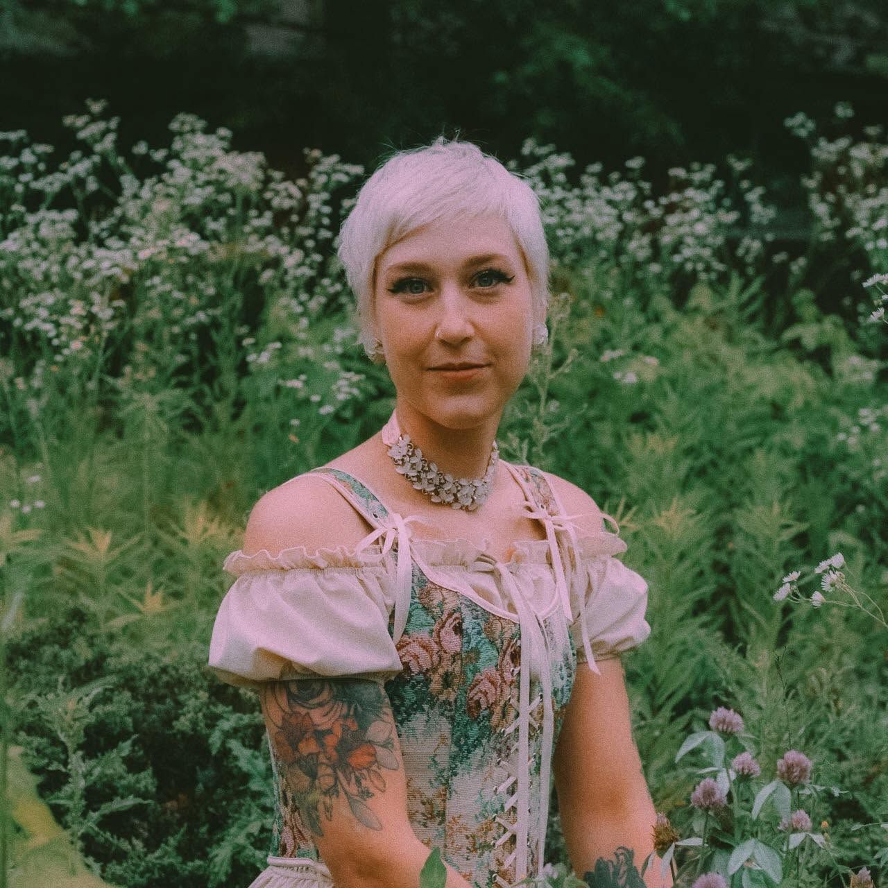 Short-haired tattooed woman in floral dress standing among wildflowers in a lush green garden.