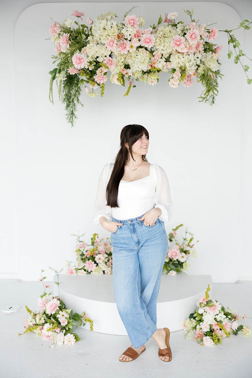 Woman in white top and jeans posing in front of pink and white floral arrangement on white backdrop.