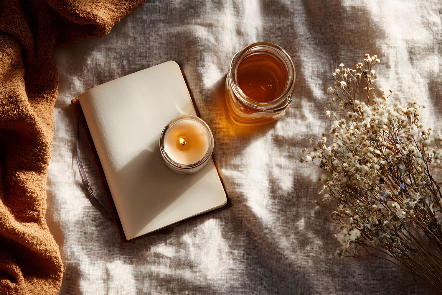 Cozy flat lay with open journal, lit candle, honey jar, and dried flowers on linen fabric in warm sunlight.