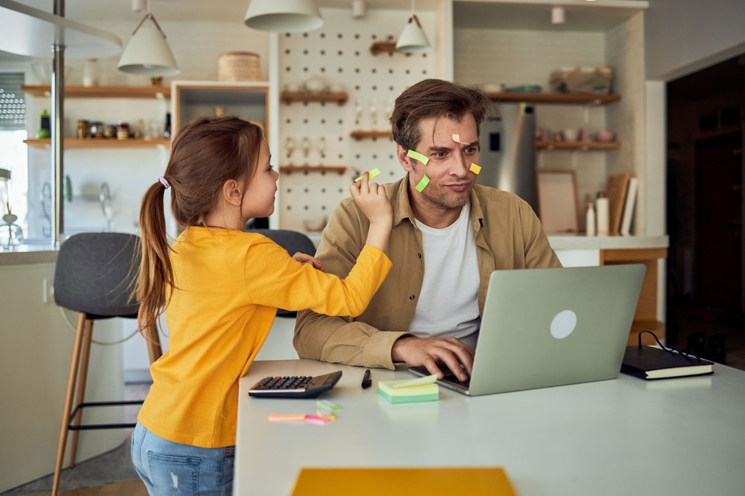 Young girl placing sticky notes on her father's face while he works on a laptop at home.