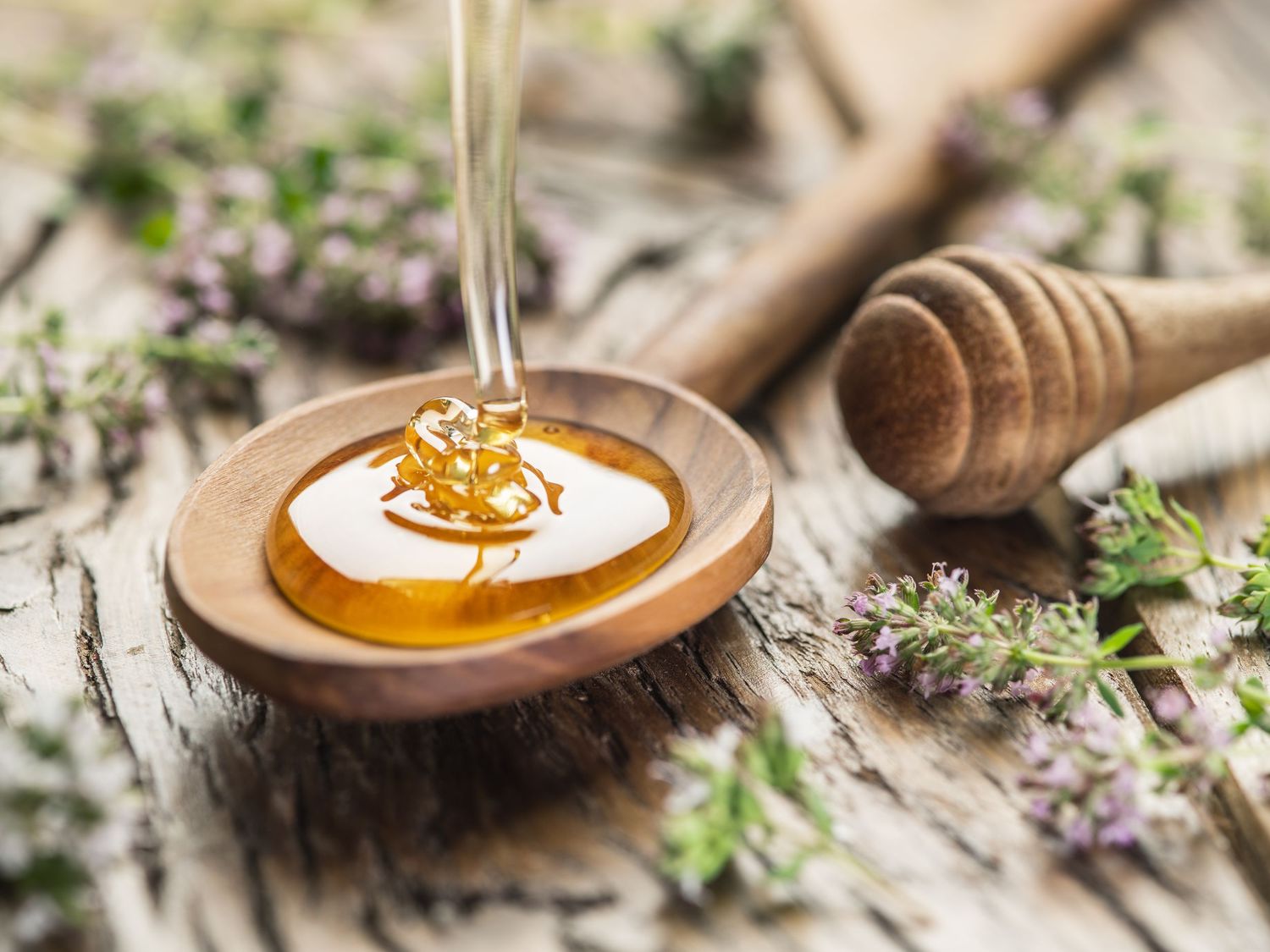 Honey dripping onto a wooden spoon surrounded by fresh thyme flowers on a rustic surface.
