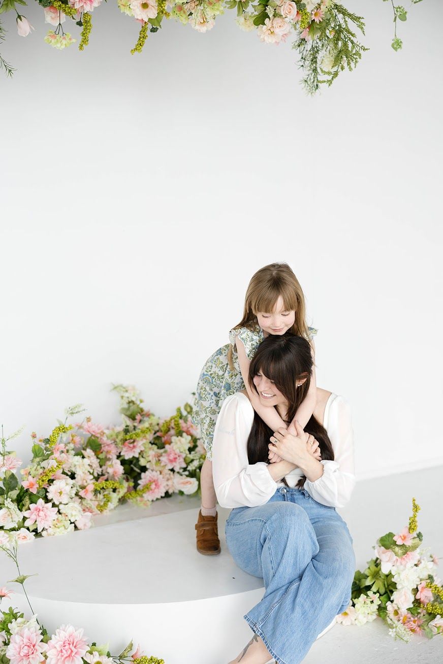 Two women embracing in a floral studio setting with pink and white flowers.