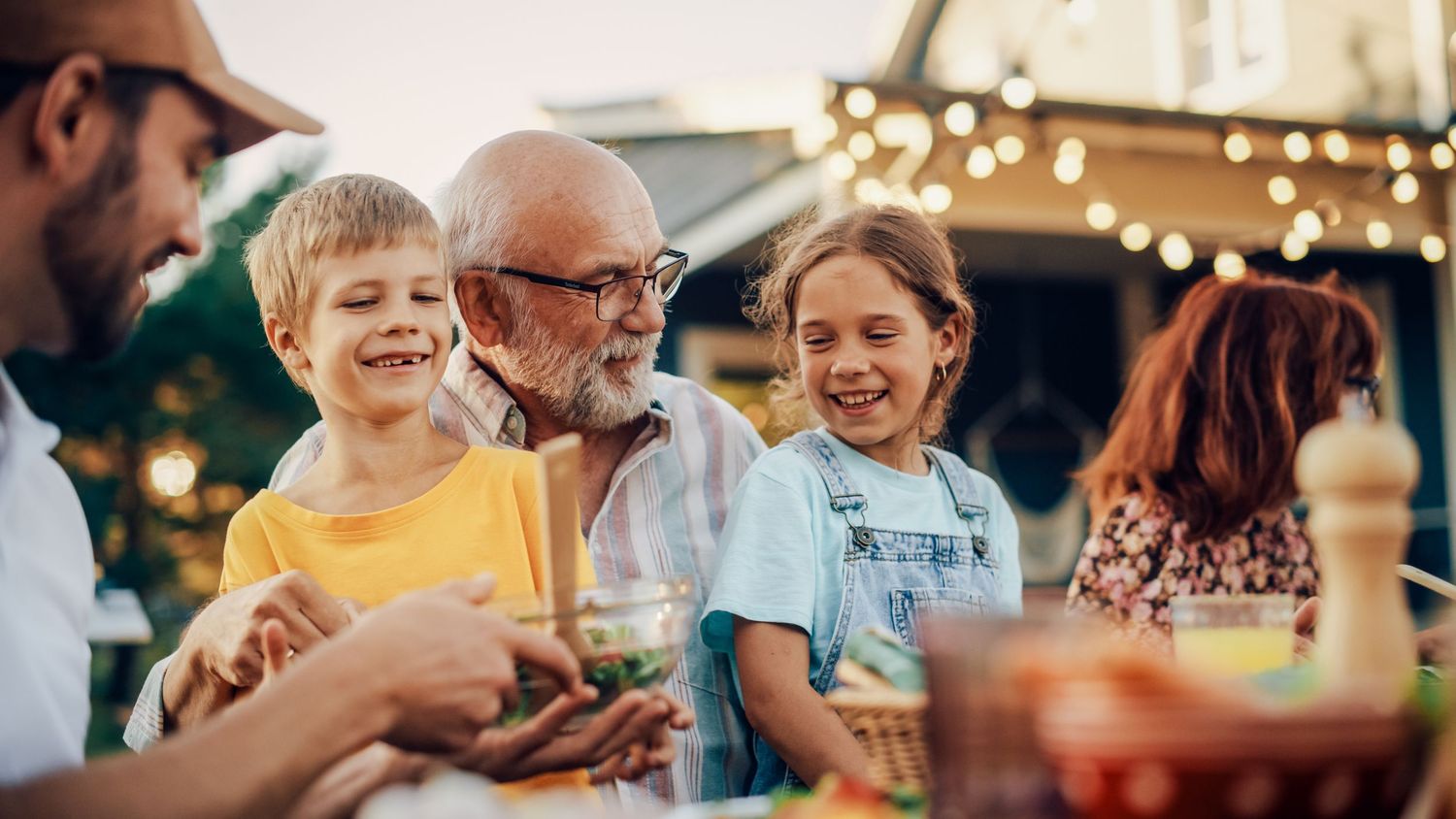 Multi-generational family laughing together at an outdoor dinner party with string lights in the background.