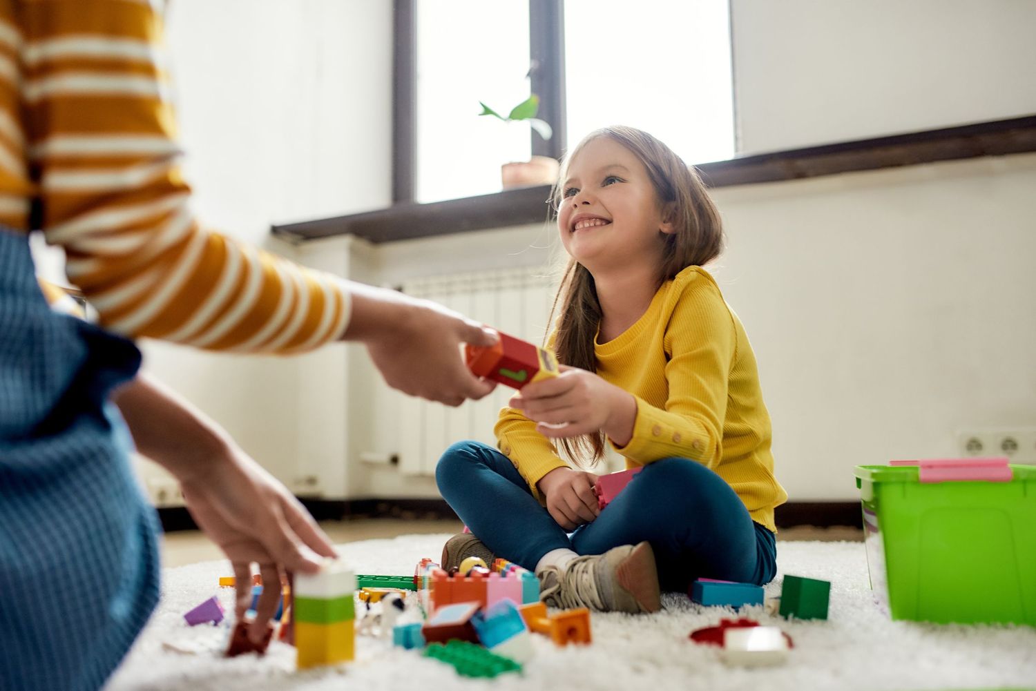 Young girl smiling while playing with colorful building blocks on a carpet with an adult indoors.