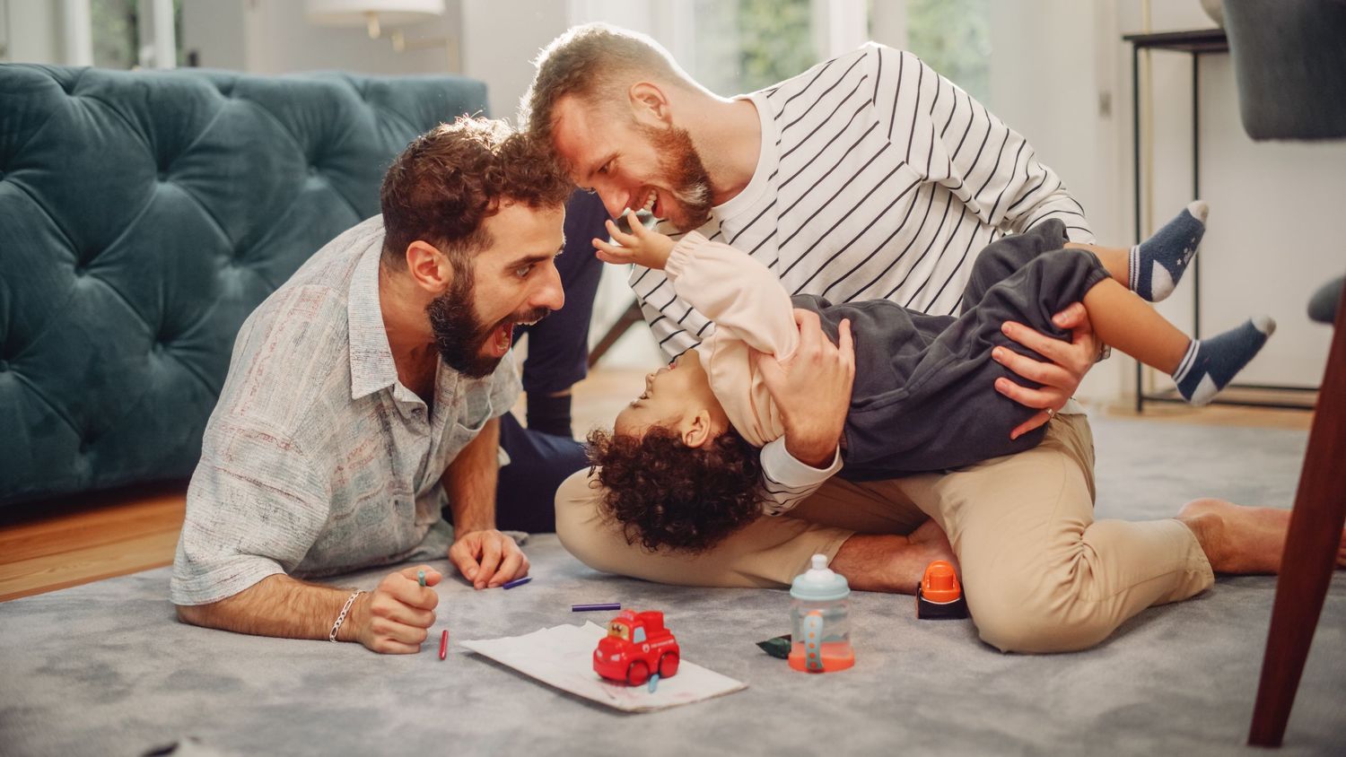 Two dads playing with their young child on living room floor, laughing and bonding together at home.