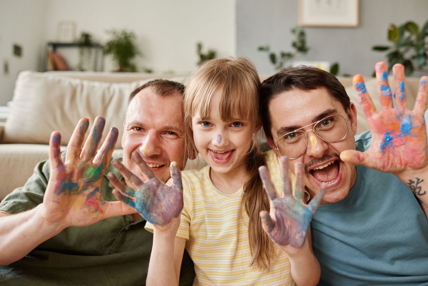 Two men and a girl smile showing colorful paint-covered hands indoors.