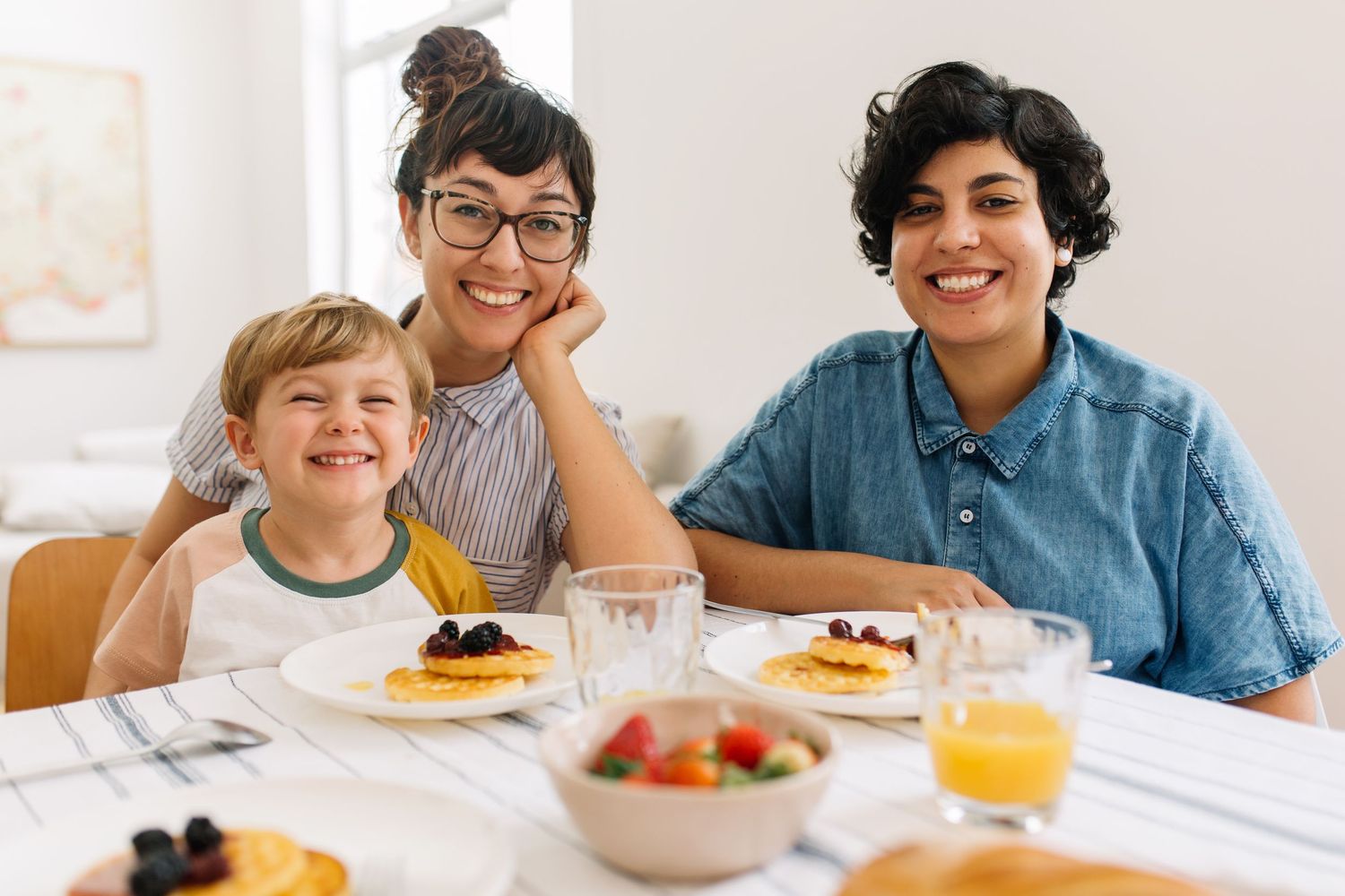 Two smiling mothers and young son enjoying a healthy breakfast with pancakes and fruit at home.