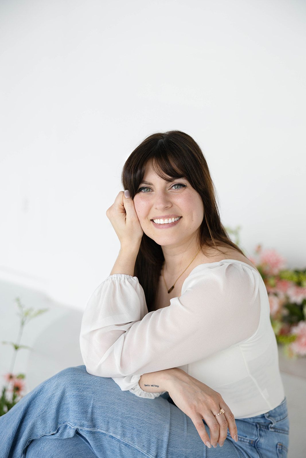 Smiling brunette woman with blue eyes wearing a white blouse and jeans, sitting in a bright airy space with pink flowers.