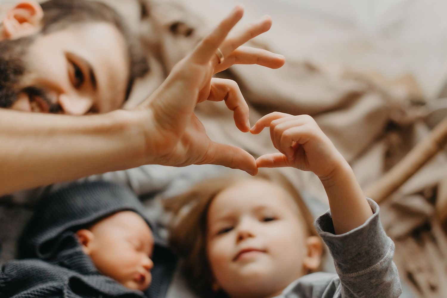 Father and child making heart shape with hands while lying with newborn baby on bed.