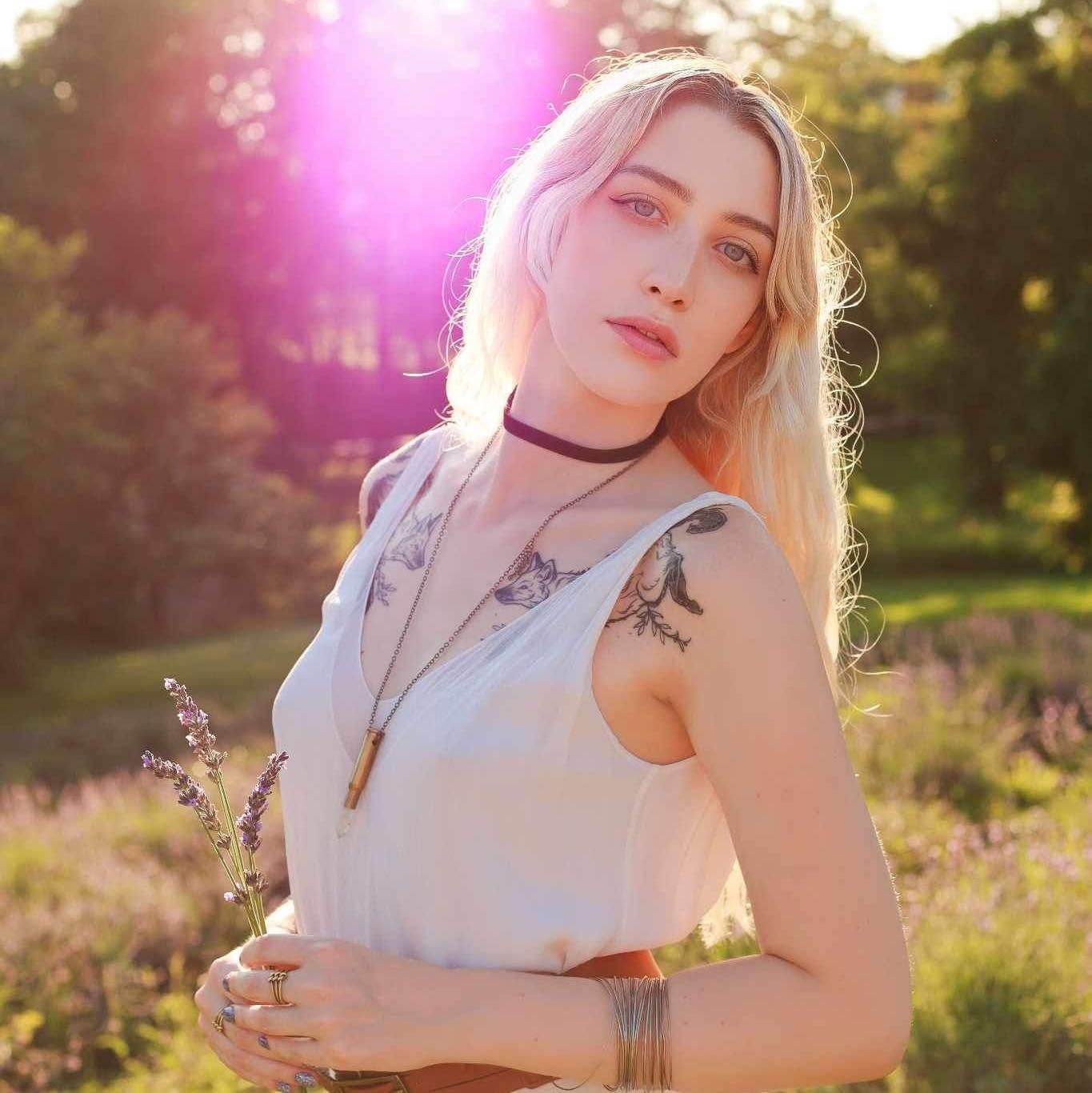 Tattooed blonde woman in white dress holding lavender in a sunlit garden at golden hour.