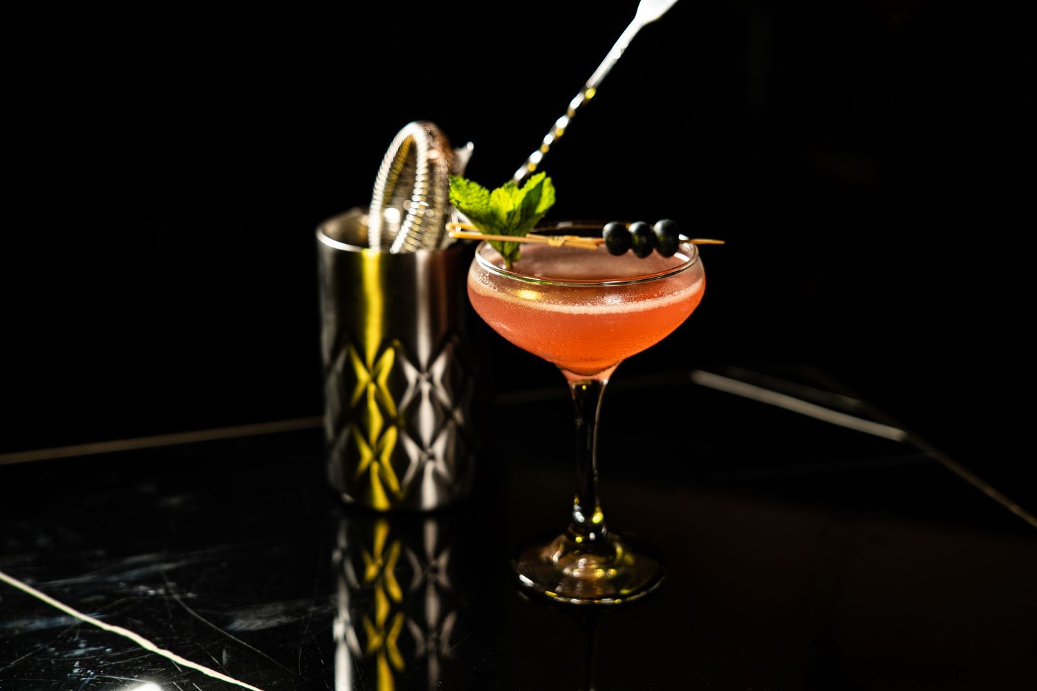 Two craft cocktails with garnishes on a black surface, one red in a coupe glass and one in a metallic shaker.