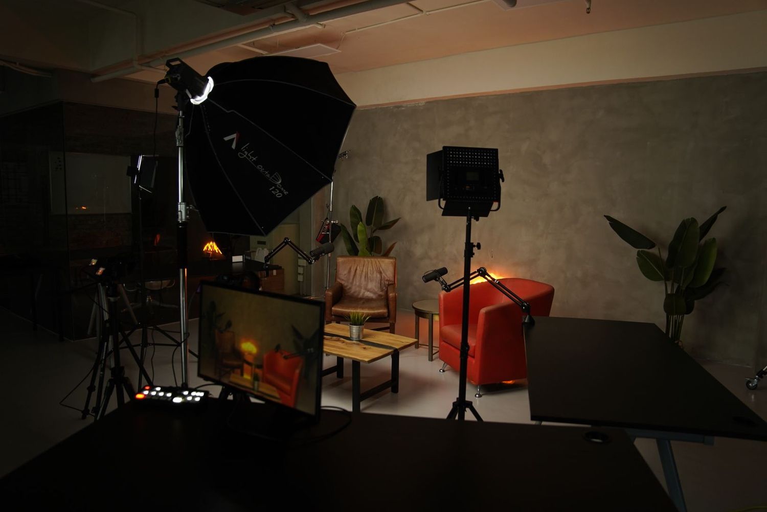 Professional photo studio setup with lighting equipment, umbrella softbox, tripod, and orange chair against a gray wall.